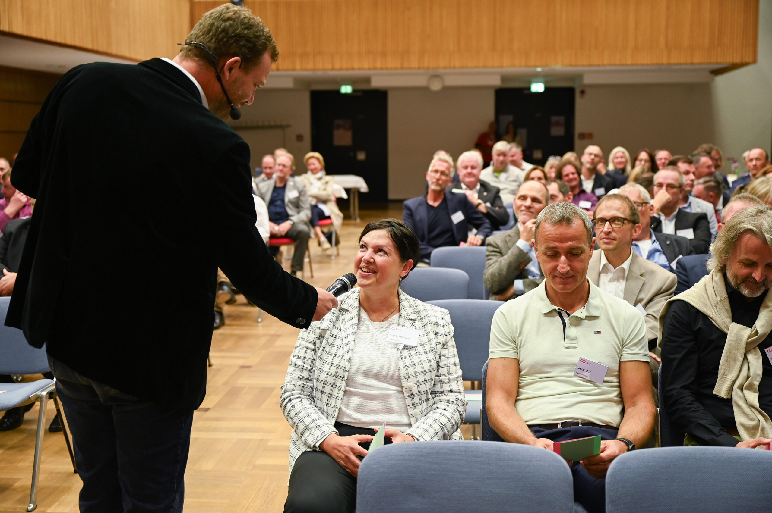 Ein Redner in einem schwarzen Jackett, der die Hochschule Coburg vertritt, unterhält sich in einem Konferenzraum mit einem Publikum. Er hält einer sitzenden Frau in einem weißkarierten Blazer, die lächelt, ein Mikrofon hin. Das aufmerksame Publikum sitzt in Reihen und wirkt engagiert.