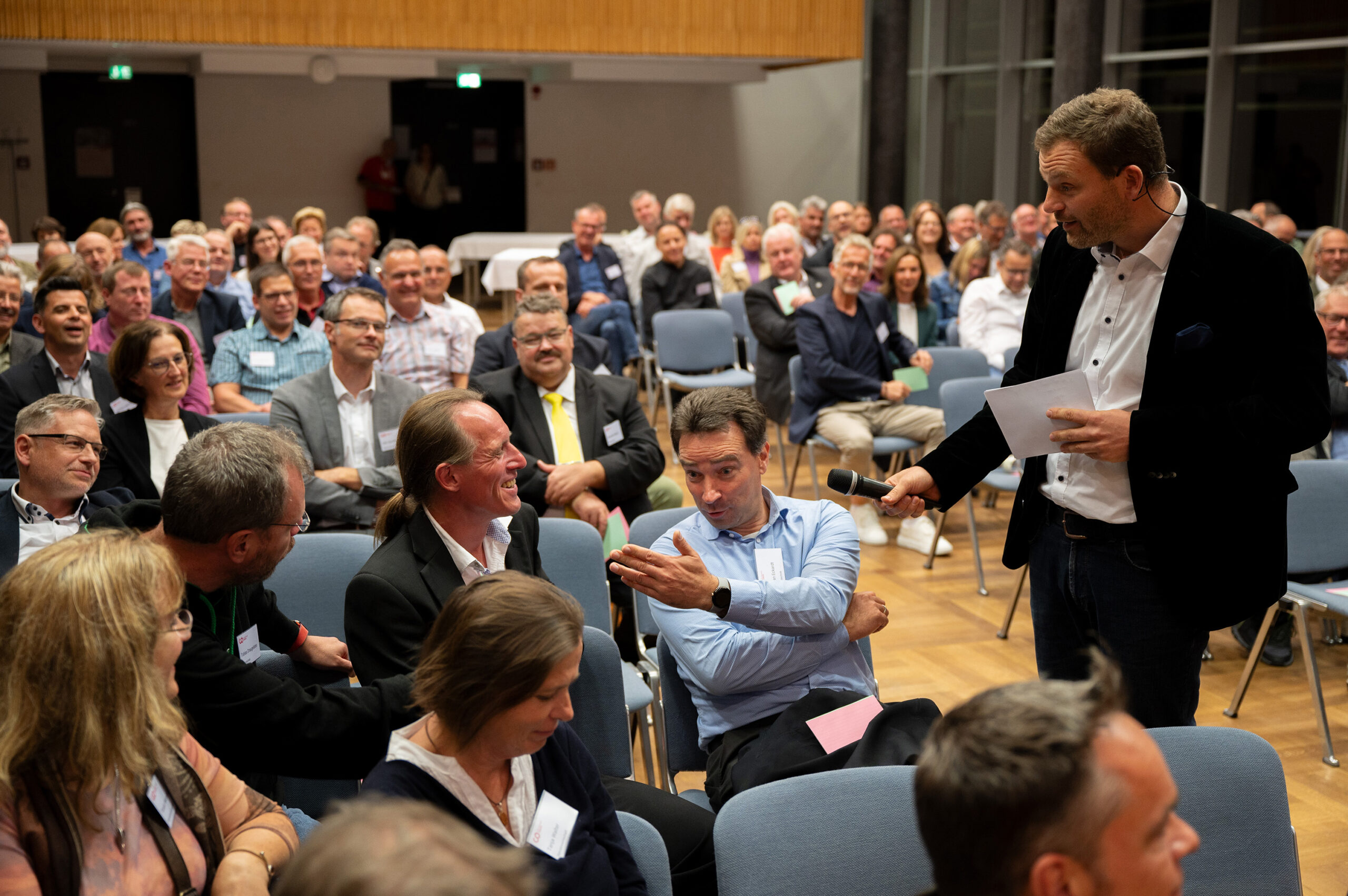 Ein Mann mit einem Mikrofon steht inmitten eines sitzenden Publikums an der Hochschule Coburg und spricht mit einem Mann, der lebhaft gestikuliert. Das Publikum, das in Reihen im hell erleuchteten Hörsaal sitzt, beobachtet ihre Interaktion mit großem Interesse.