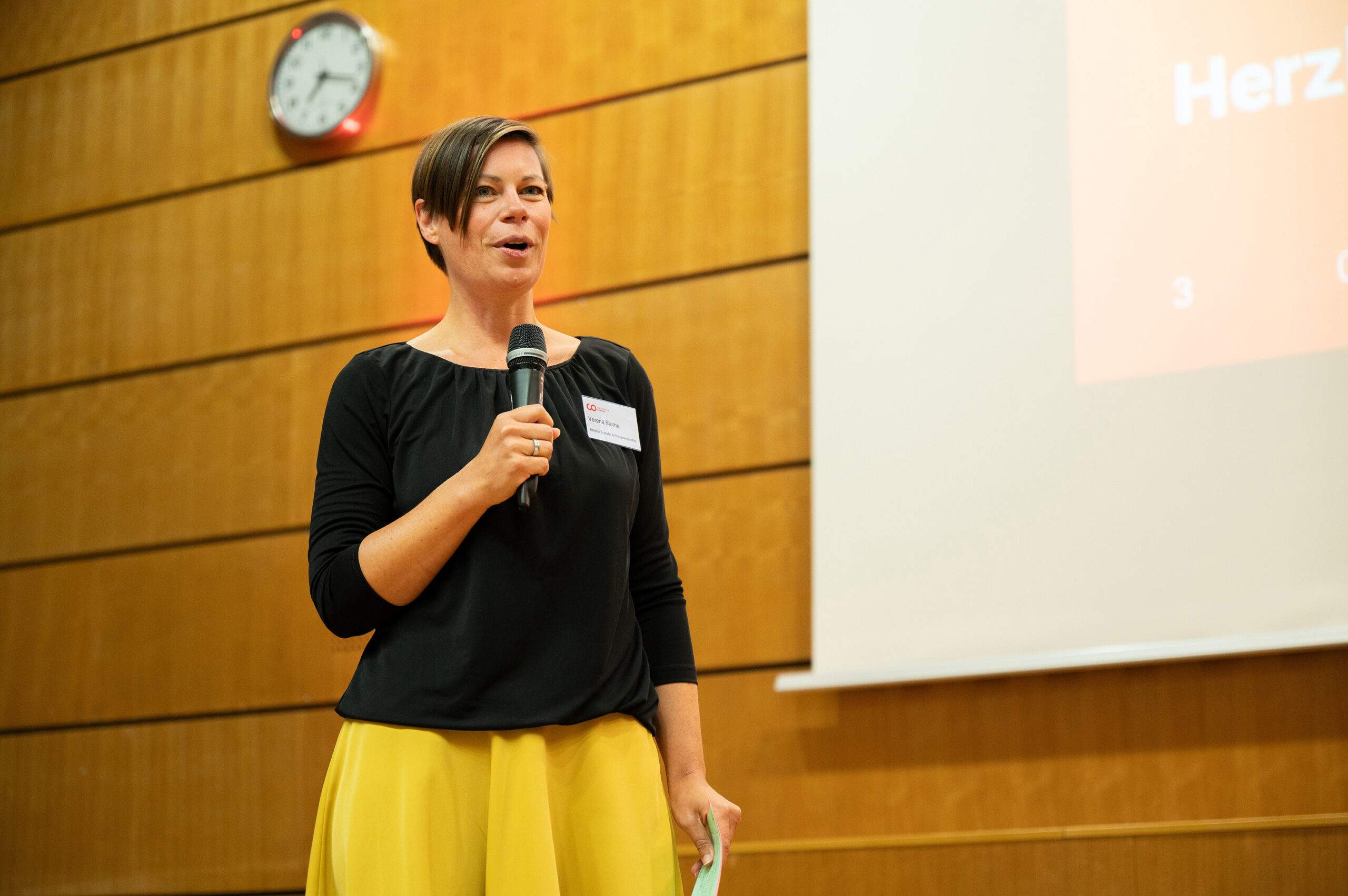 In einem Raum mit Holzwänden der Hochschule Coburg spricht eine Person mit einem Mikrofon. An der Wand ist eine Uhr zu sehen, rechts davon ist eine Projektionsleinwand. Die Rednerin, die ein schwarzes Oberteil und einen gelben Rock trägt, zieht das Publikum in ihren Bann.