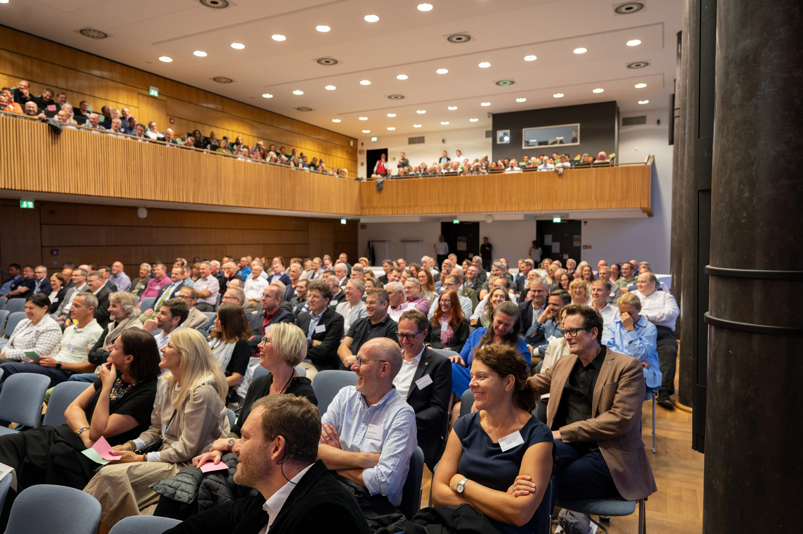 Im Hörsaal der Hochschule Coburg sitzen zahlreiche Zuhörer und hören aufmerksam einem Vortragenden zu. Der Saal ist mit Menschen unterschiedlichen Alters gefüllt, die Wände sind mit Holz verkleidet, und auf dem Balkon sitzen weitere Zuhörer.