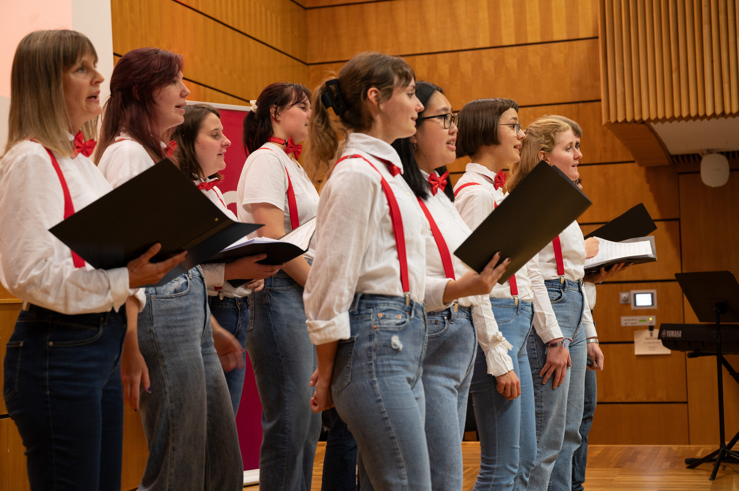 In einem beeindruckenden holzgetäfelten Raum der Hochschule Coburg singt ein Chor in weißen Hemden, roten Hosenträgern und Jeans harmonisch. Jedes Mitglied hält einen schwarzen Ordner mit Noten in der Hand, während das Keyboard seinen Auftritt sanft begleitet.