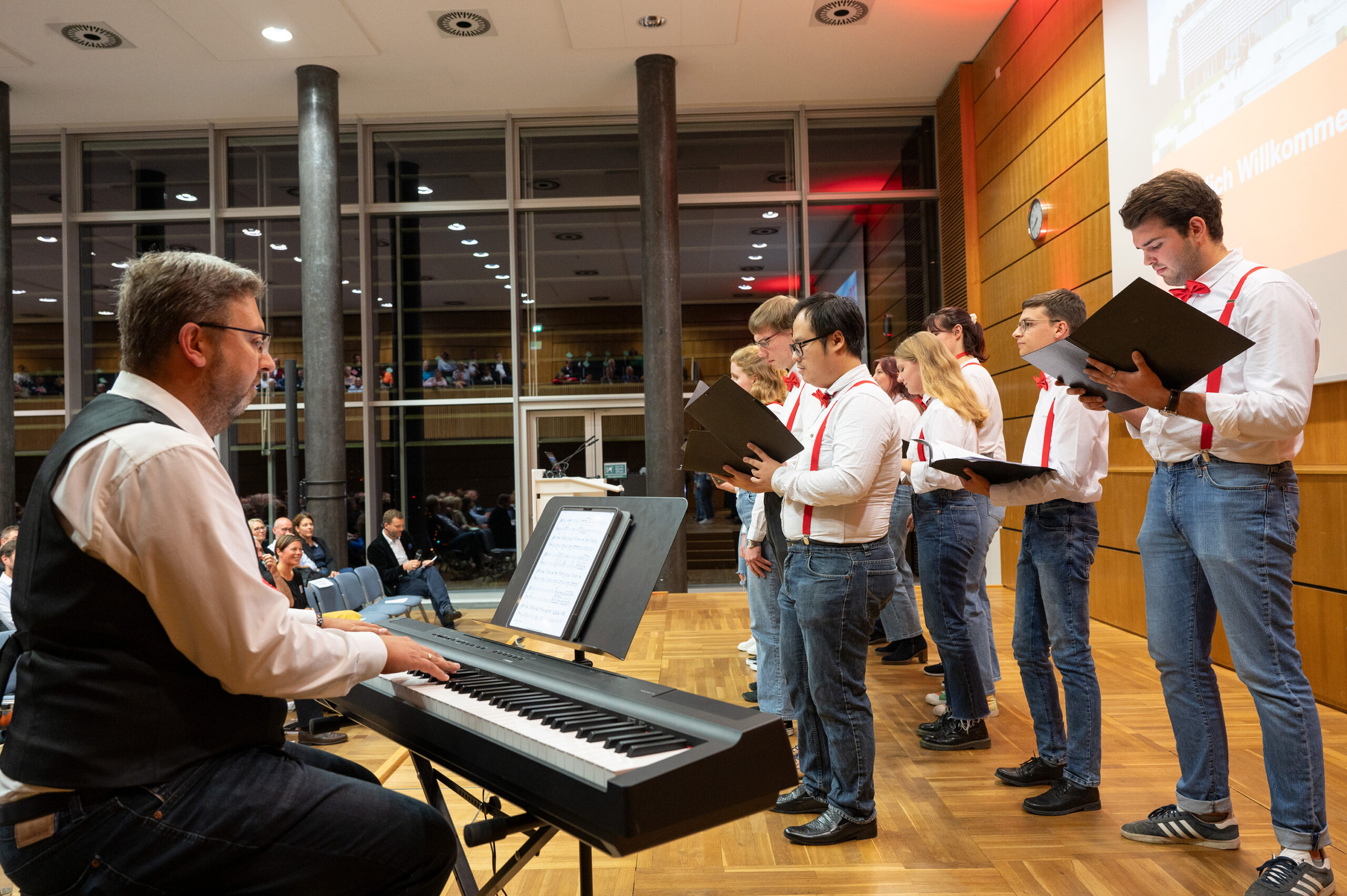 Eine Gruppe von Chorsängern der Hochschule Coburg, in weißen Hemden und roten Fliegen, tritt in einem modernen Konzertsaal auf. Sie stehen in einer Reihe und halten schwarze Mappen mit Notenblättern in der Hand, während ein Mann in der Nähe auf einem elektronischen Keyboard spielt. Im Hintergrund sitzt das Publikum.