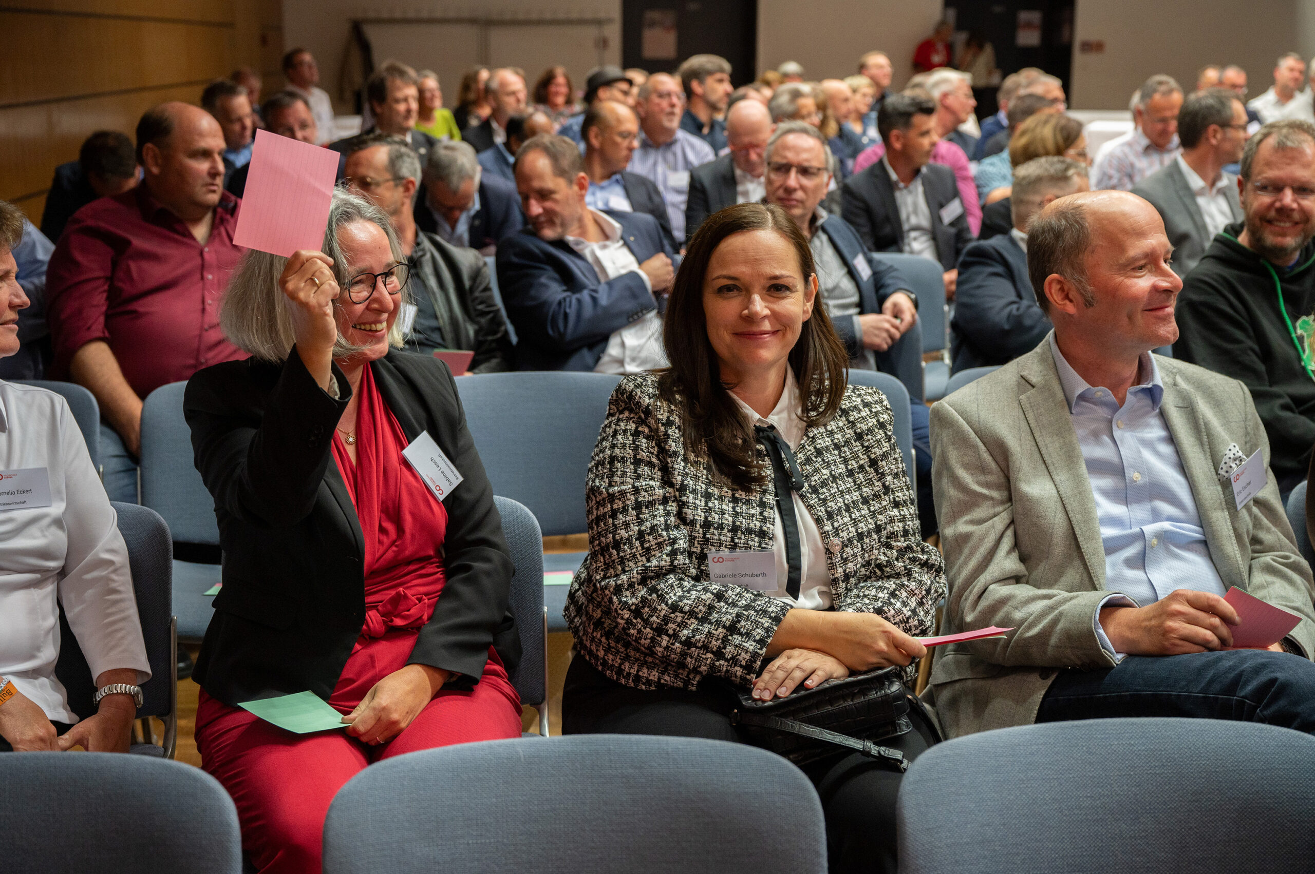 In einem überfüllten Konferenzraum der Hochschule Coburg sitzen die Teilnehmer. Eine Frau in der ersten Reihe hält lächelnd eine rosa Karte hoch. Die anderen Teilnehmer sind auf eine Präsentation oder Diskussion konzentriert. Die Leute tragen Business-Kleidung, was die professionelle Atmosphäre der Hochschule widerspiegelt.