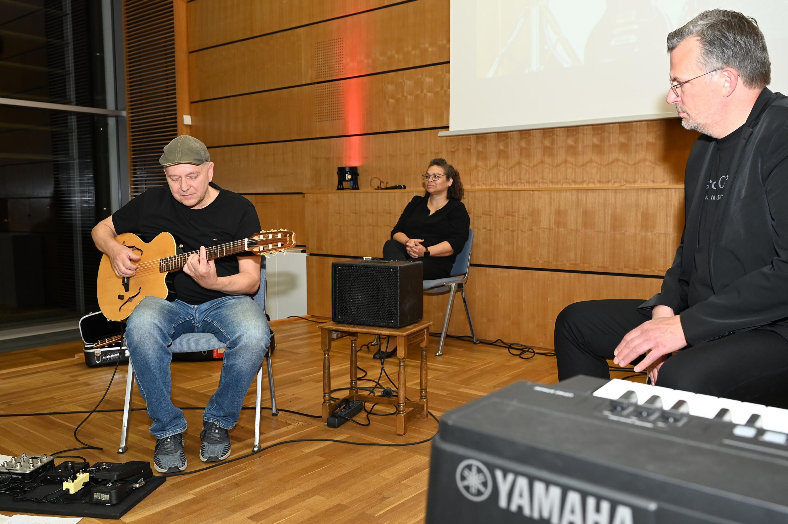 Ein Musiker mit Mütze spielt im Sitzen eine elektrische Gitarre, im Hintergrund sitzt eine Person und eine weitere neben einem Yamaha-Keyboard. Alle sind in einem holzgetäfelten Raum versammelt, der an das gemütliche Ambiente der Hochschule Coburg erinnert.