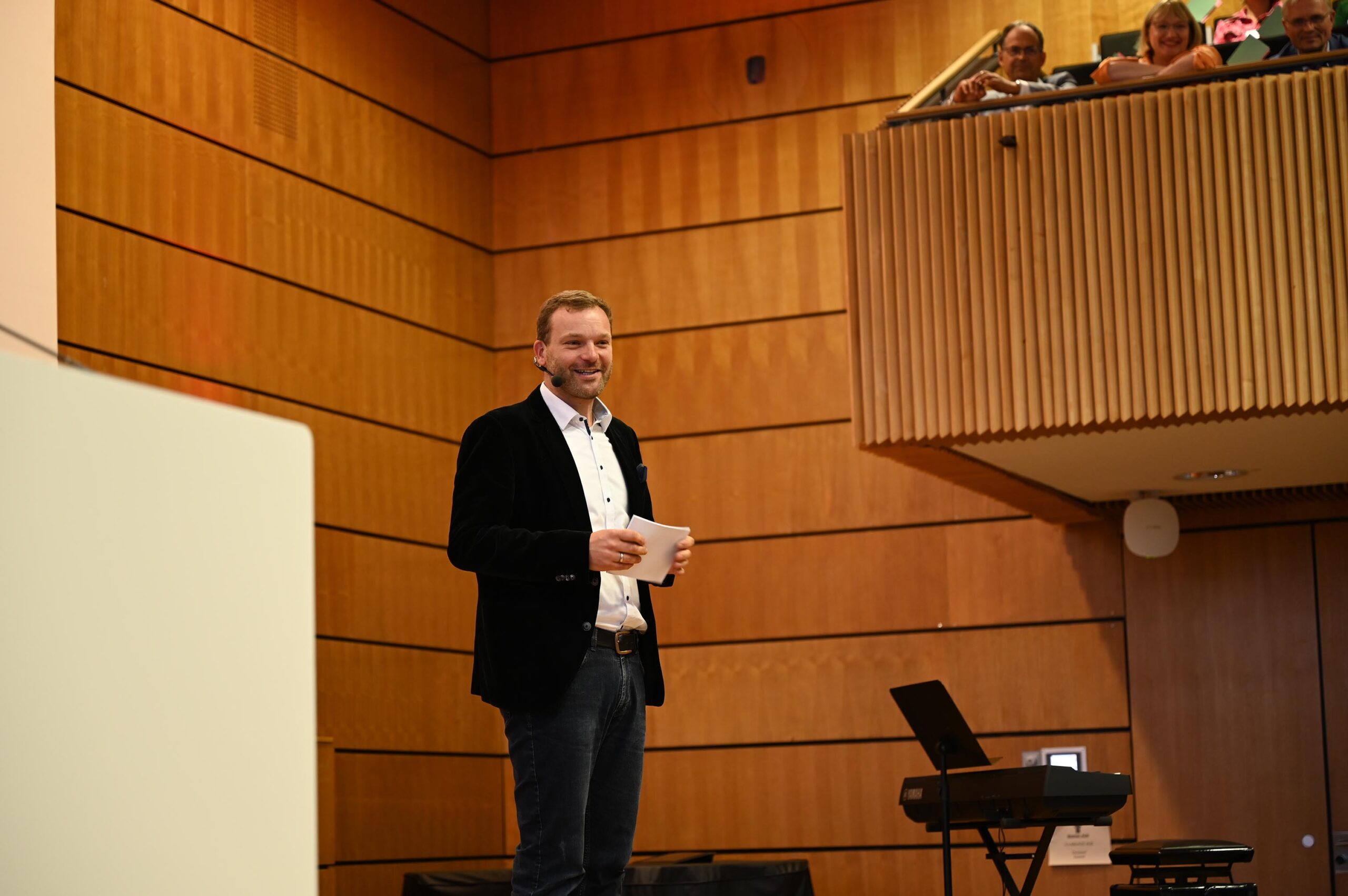 Ein Mann in einem dunklen Blazer und Jeans spricht auf der Bühne der Hochschule Coburg und hält Papiere in der Hand. Die Kulisse scheint ein hölzerner Hörsaal zu sein, über dem eine Gruppe von Menschen von einem Balkon aus zusieht. Auf der rechten Seite der Bühne ist ein Klavier zu sehen.