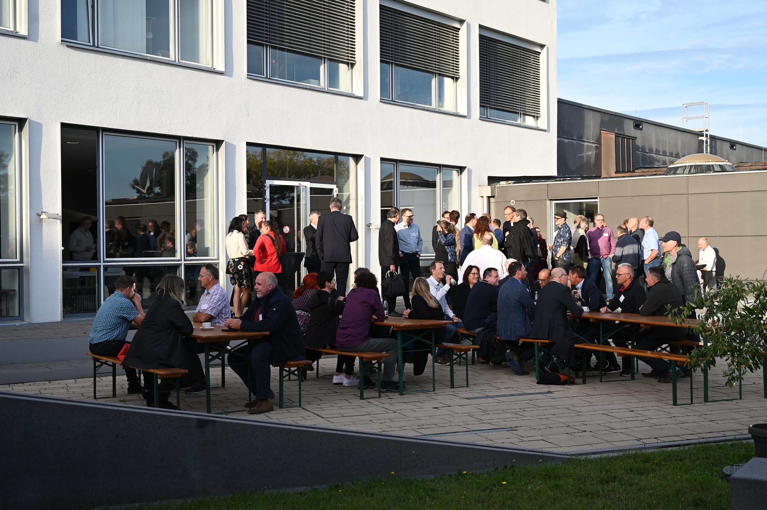 Eine Gruppe von Menschen hat sich vor einem Gebäude der Hochschule Coburg versammelt. Einige stehen im Gespräch, andere sitzen an Tischen und Bänken. Die Atmosphäre scheint eine gesellige Veranstaltung oder ein Networking-Event an einem sonnigen Tag zu sein.