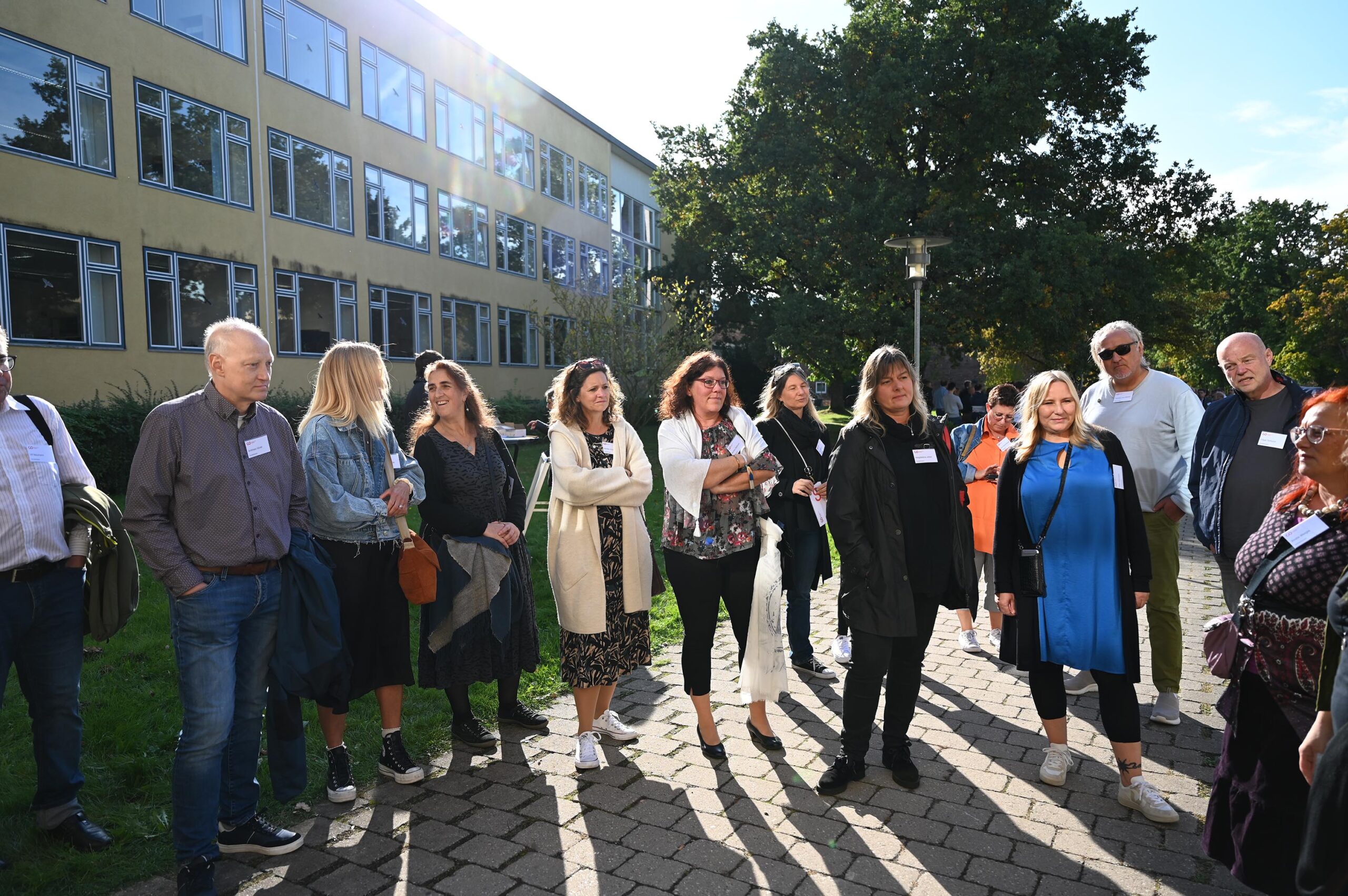 An einem sonnigen Tag steht eine Gruppe von Menschen draußen in der Nähe eines Gebäudes mit großen Fenstern der Hochschule Coburg. Einige tragen Namensschilder. Sie haben sich auf einem gepflasterten Weg versammelt, umgeben von Bäumen und Grün, und unterhalten sich.