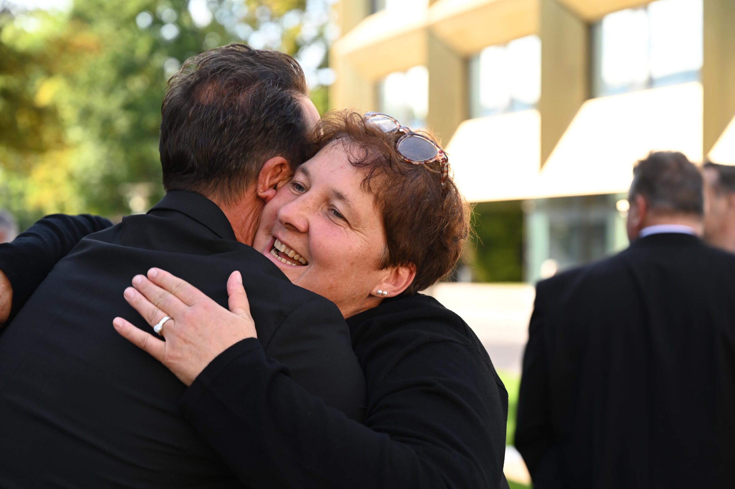 Eine Frau mit Brille auf dem Kopf umarmt freudig einen Mann im Freien in einer sonnigen Umgebung in der Nähe der Hochschule Coburg. Beide sind dunkel gekleidet und strahlen Freude aus. Im Hintergrund sind zwei weitere Personen leicht unscharf zu sehen.