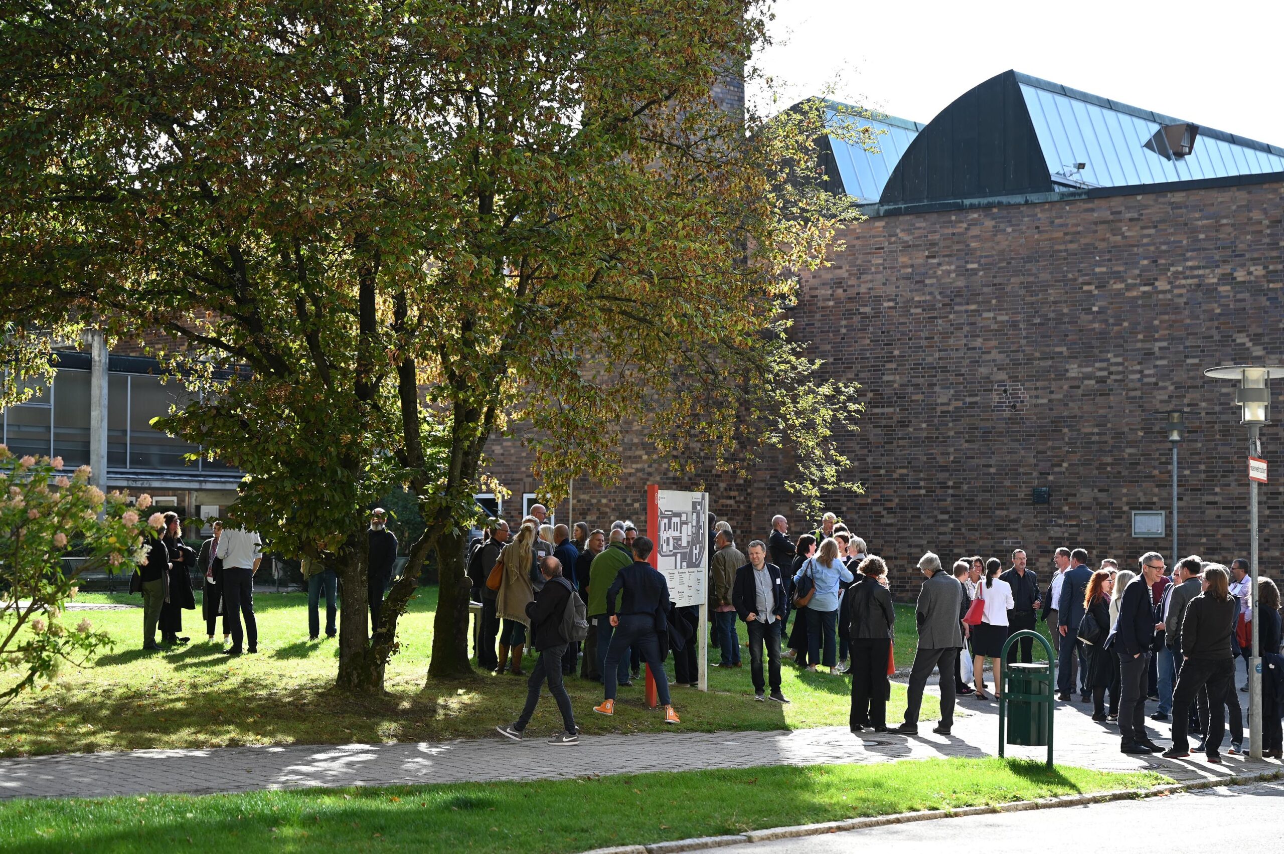 An einem sonnigen Tag steht eine Gruppe von Menschen auf einer Wiese in der Nähe des großen Backsteingebäudes der Hochschule Coburg. Einige halten Schilder hoch, von denen eines deutlich zu sehen ist, während andere sich unter einem belaubten Baum unterhalten. Im Hintergrund sind moderne Architektur und ein klarer Himmel zu sehen.