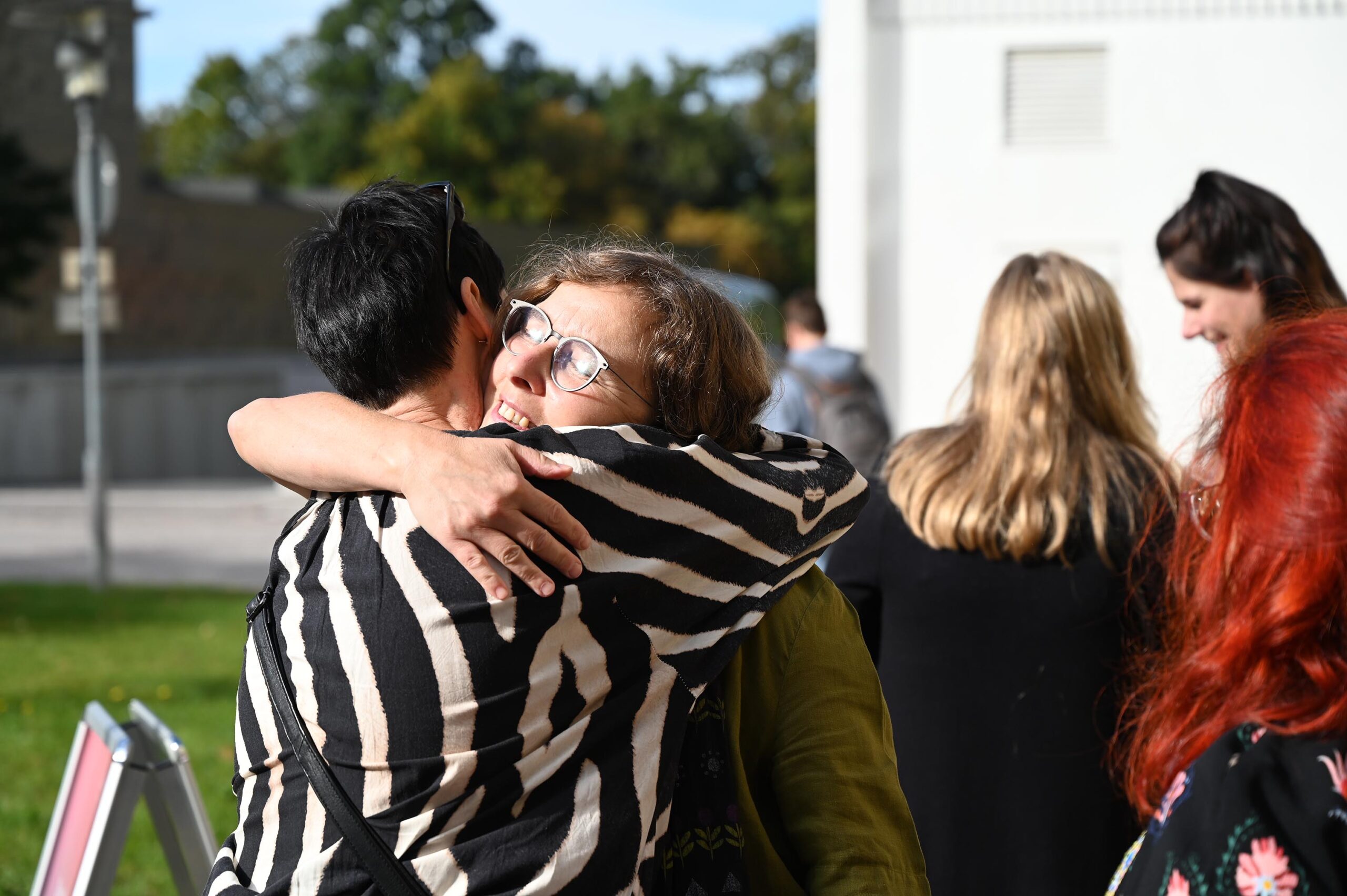 Zwei Menschen umarmen sich herzlich an einem sonnigen Tag im Freien an der Hochschule Coburg. Einer trägt ein Hemd mit Zebramuster, der andere hat eine Brille und eine grüne Jacke. In der Nähe versammeln sich mehrere andere auf dem grasbewachsenen Campus mit Gebäuden im Hintergrund.