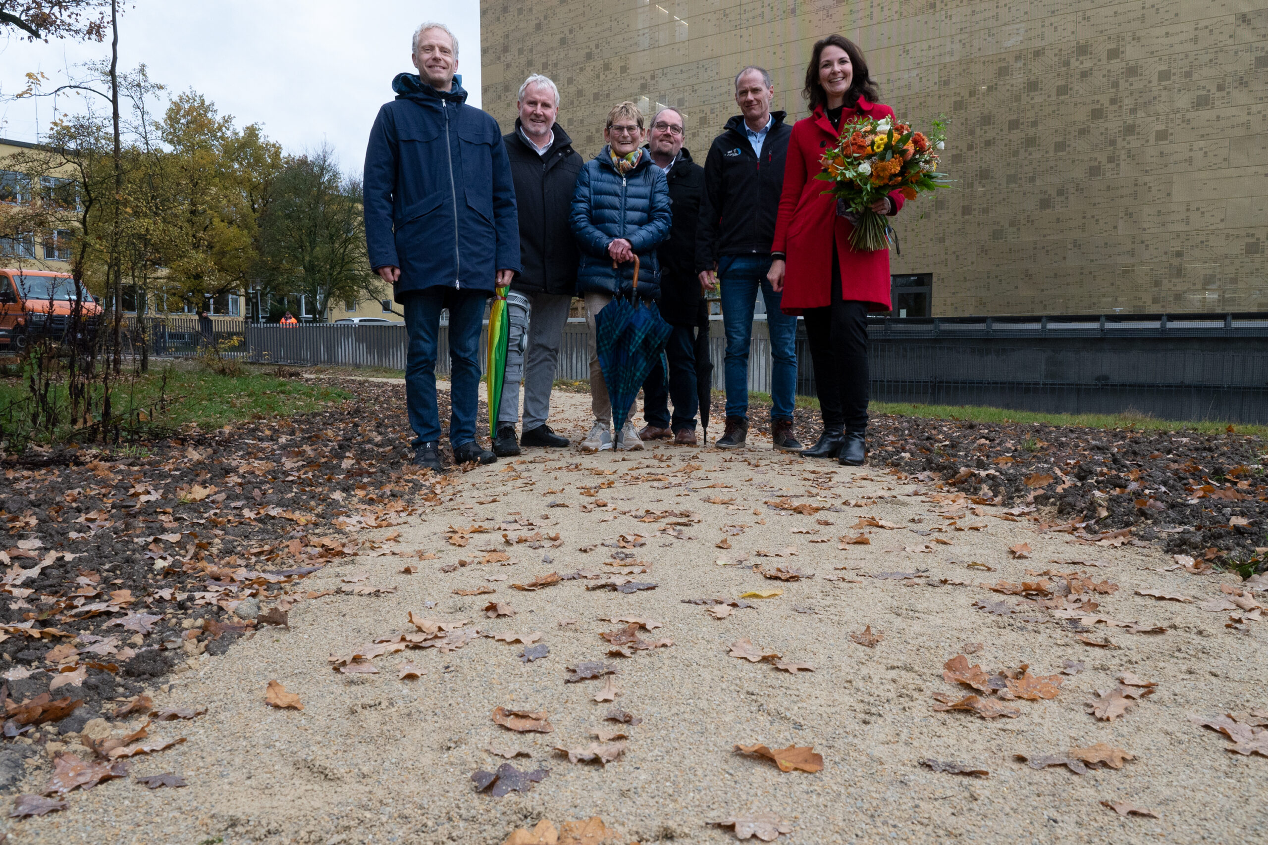 Eine Gruppe von sechs Personen steht auf einem mit Herbstlaub geschmückten Kiesweg, einer hält einen Blumenstrauß in der Hand. Sie befinden sich im Freien in der Nähe der Hochschule Coburg, hinter ihnen ein großes Gebäude und an den Seiten Bäume.