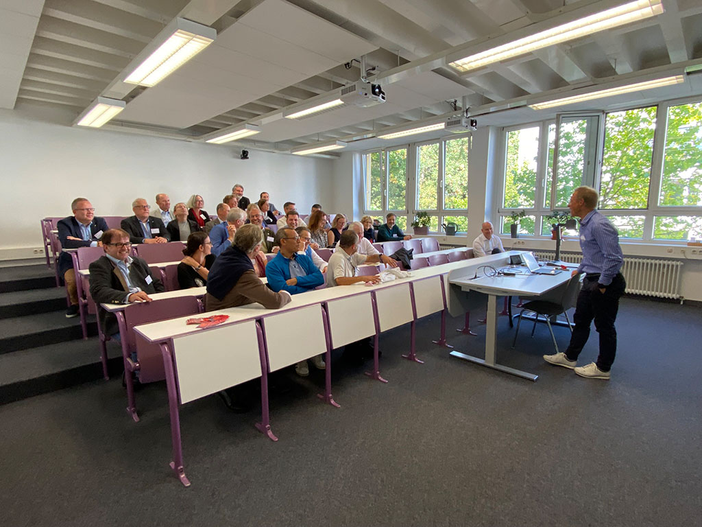 Ein Mann hält in einem Hörsaal mit violetten Stühlen an der Hochschule Coburg einen Vortrag. Der Raum mit den großen Fenstern und Deckenbeleuchtung ist voller aufmerksamer Zuhörer, von denen einige fleißig Notizen machen.