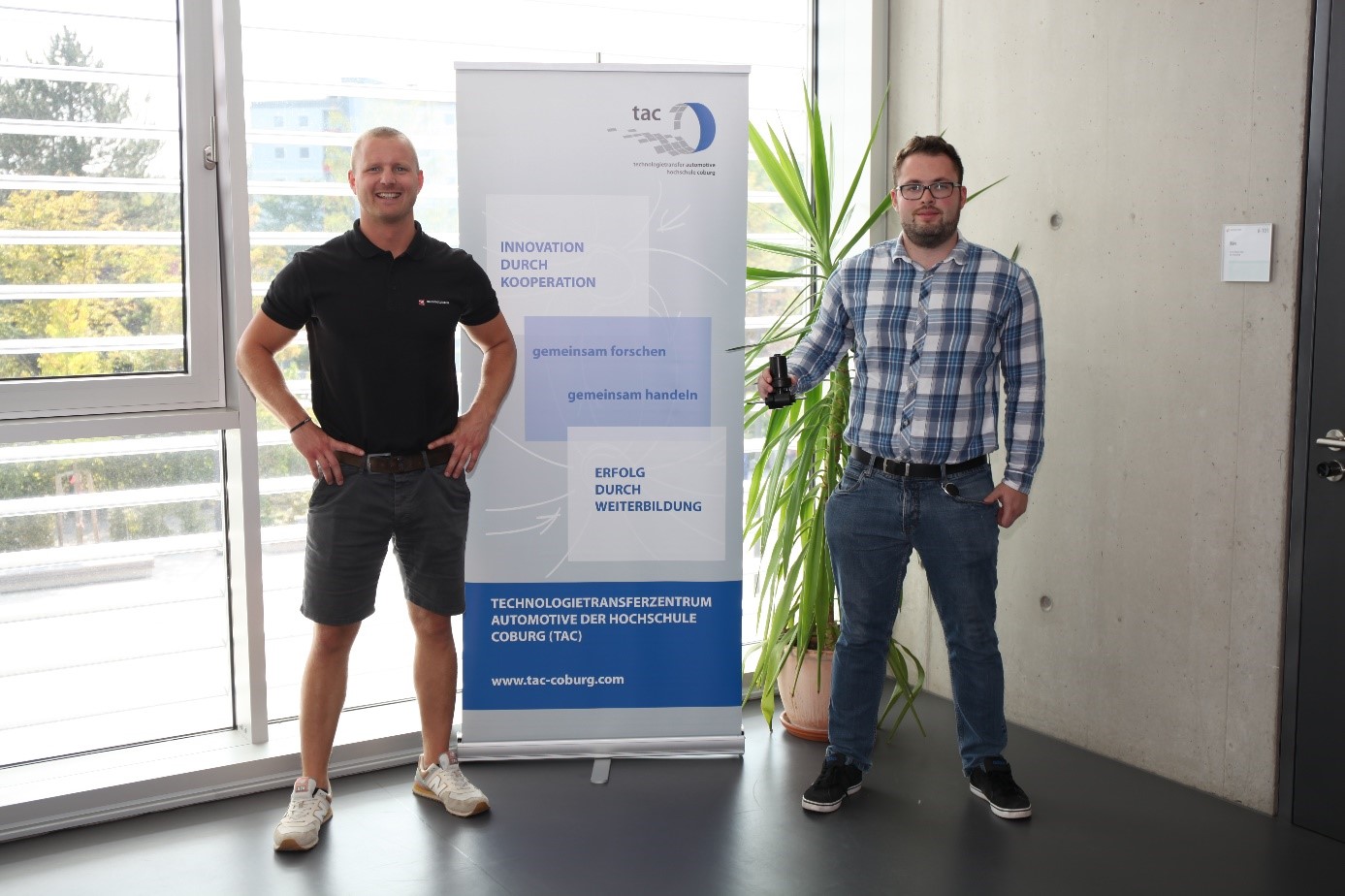 Zwei Männer stehen drinnen neben einem Roll-up-Banner und verkörpern damit den Innovationsgeist und die Zusammenarbeit mit der Hochschule Coburg. Der Mann links trägt ein schwarzes Poloshirt und Shorts, während sein Gegenüber sich für ein kariertes Hemd und Jeans entscheidet.