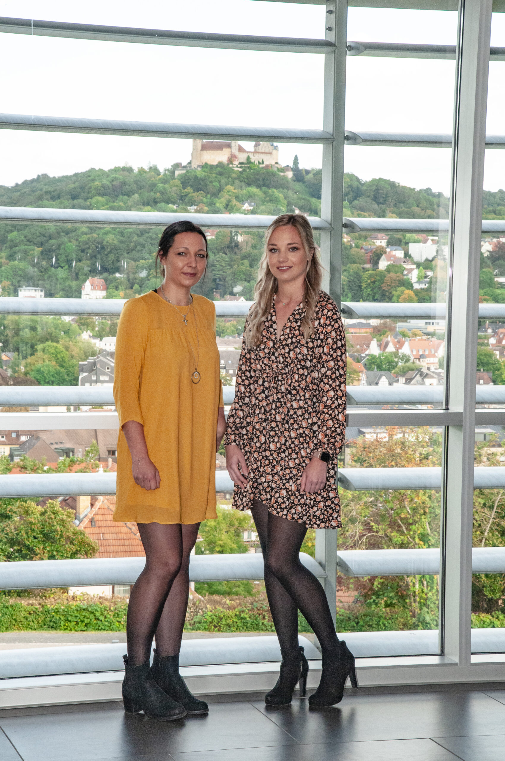 Zwei Frauen stehen zusammen vor einem großen Fenster mit Blick auf einen üppigen, grünen Hügel, auf dem sich ein Schloss in der Nähe der Hochschule Coburg befindet. Eine trägt ein gelbes Kleid, die andere ein geblümtes Kleid. Im Hintergrund sind die Stadtlandschaft und Bäume zu sehen.