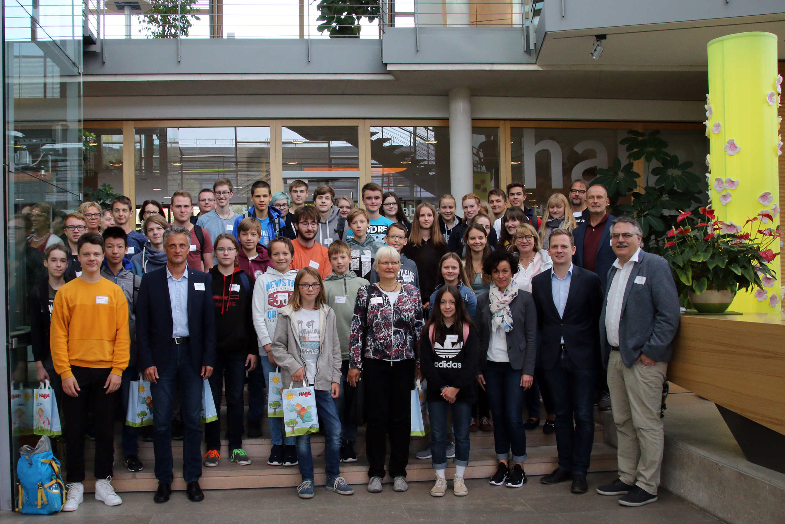 Eine große Gruppe von Menschen, darunter Erwachsene und Jugendliche, hat sich für ein Foto in einem Innenraum der Hochschule Coburg versammelt. Einige halten Geschenktüten in den Händen. Im Hintergrund ist ein modernes Interieur mit Pflanzen und Glaswänden zu sehen.