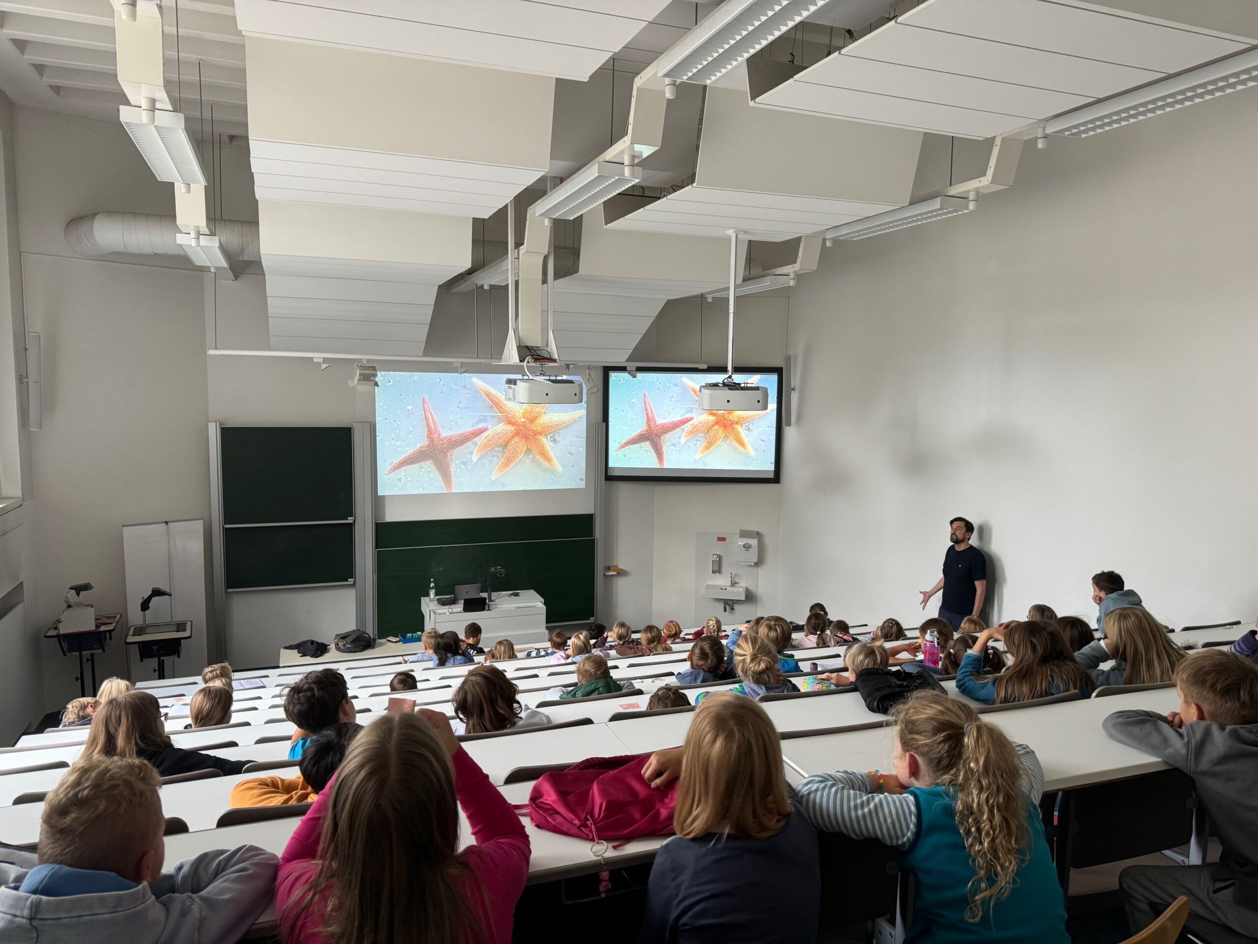 Ein Dozent der Hochschule Coburg hält einen Vortrag vor einem Klassenzimmer voller Studenten. Die Vortragsfolie zeigt Seesterne auf zwei Bildschirmen im Vordergrund. Die Studenten sitzen in abgestuften Reihen und verfolgen die Präsentation aufmerksam.