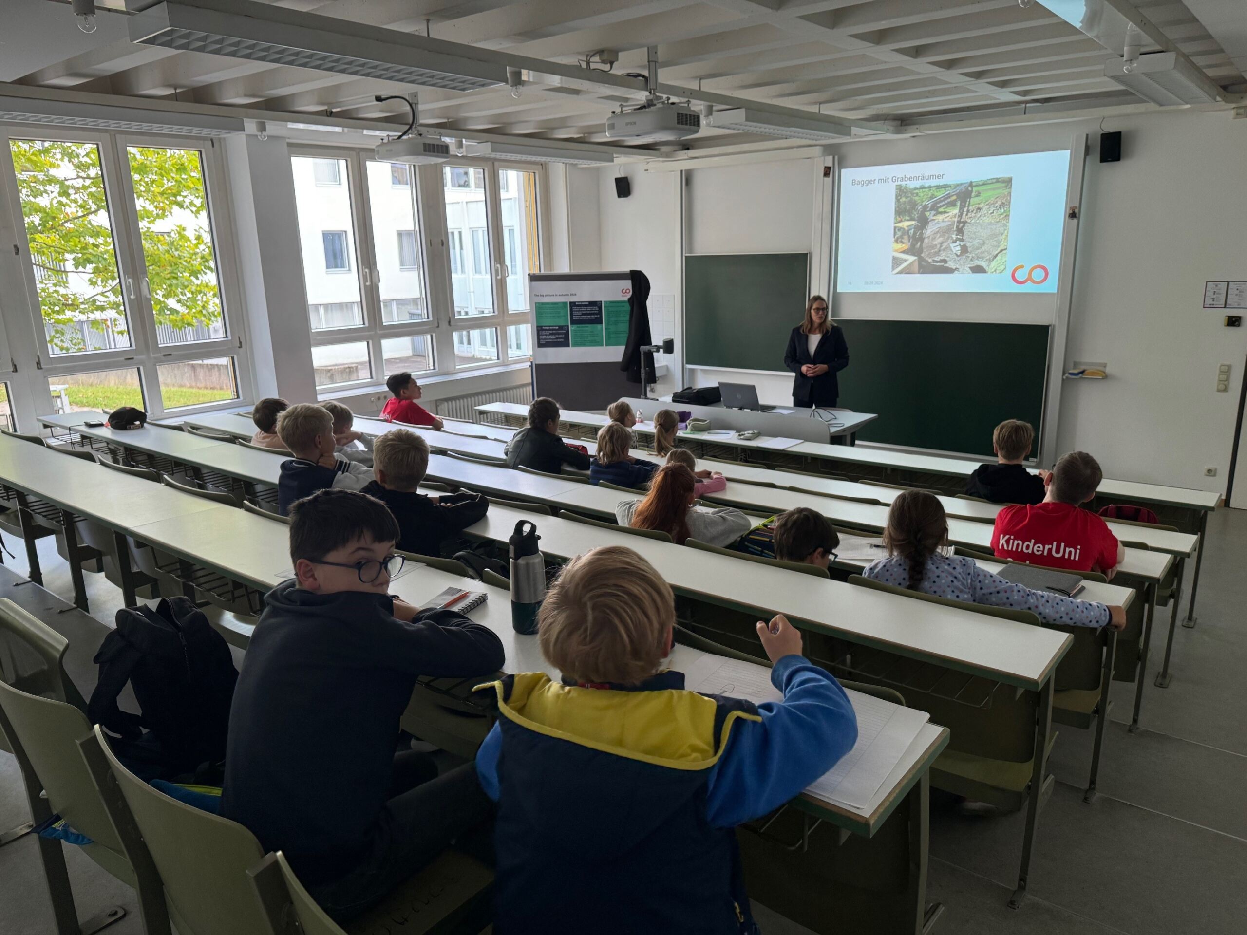 Ein Lehrer steht vor einem Hörsaal der Hochschule Coburg und präsentiert einer Gruppe von Kindern, die an ihren Schreibtischen sitzen, Folien. Der Raum ist gut beleuchtet, natürliches Licht fällt durch große Fenster an der Seite ein. Die Präsentation scheint das junge Publikum zu fesseln.