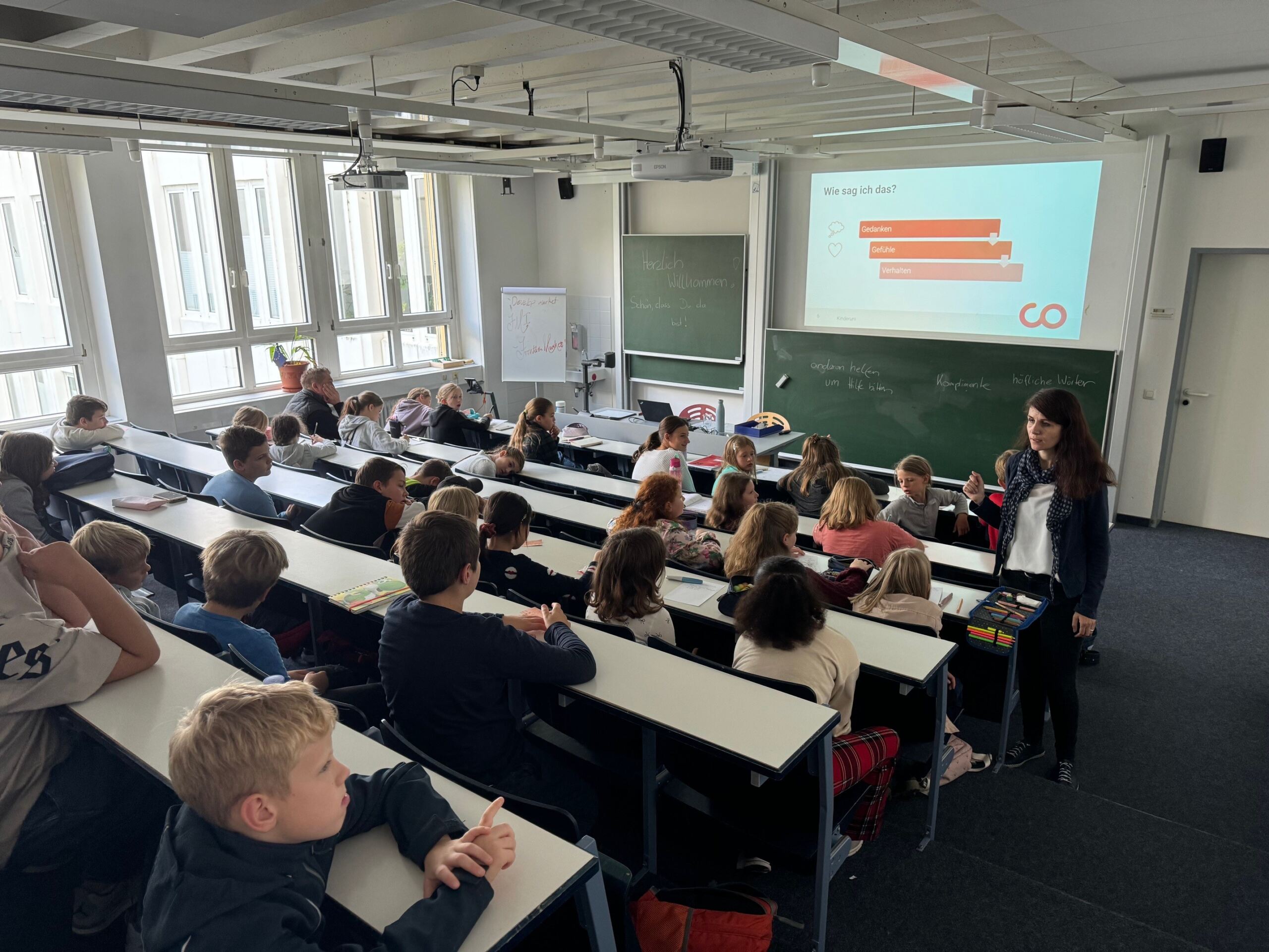 Ein Lehrer steht vor einem Klassenzimmer voller aufmerksamer, in Reihen sitzender Studierender der Hochschule Coburg. Auf einer Projektionsleinwand wird eine Präsentation mit einem Diagramm angezeigt. Das Klassenzimmer ist gut beleuchtet, natürliches Licht fällt durch große Fenster.