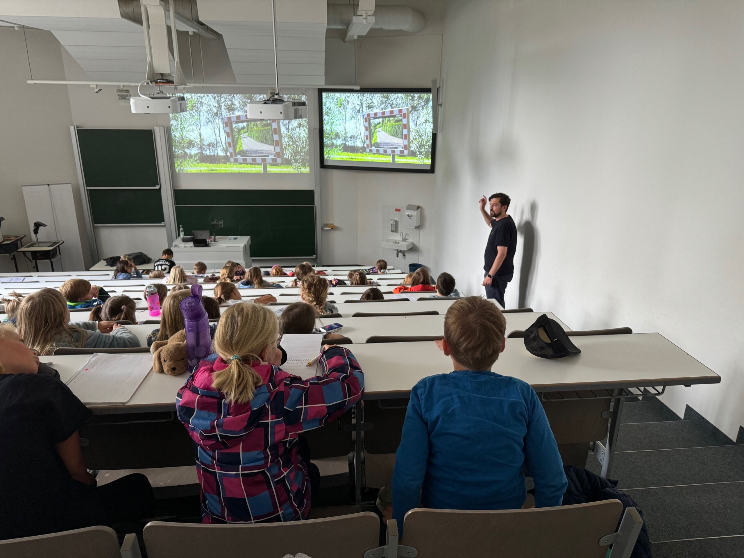 In einem modernen Hörsaal der Hochschule Coburg hält ein Dozent einen Vortrag vor Studenten, die in abgestuften Reihen sitzen, während auf zwei Bildschirmen spannende Folien mit Naturthemen laufen. Die Studenten machen sich fleißig Notizen und sind voll und ganz in das immersive Lernerlebnis vertieft.