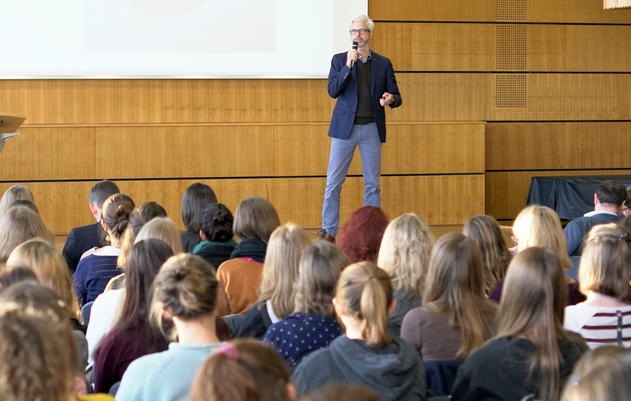 Ein Redner steht auf der Bühne der Hochschule Coburg und spricht zu einem aufmerksamen Publikum im Hörsaal. Der Raum ist mit Holzpaneelen und einer großen Projektionsleinwand ausgestattet, was die spannende Atmosphäre verstärkt, während sich die unterschiedlichsten Teilnehmer auf die Präsentation konzentrieren.