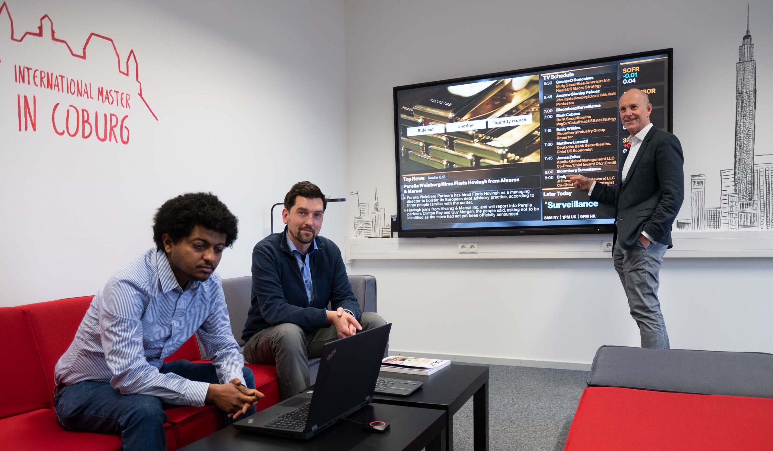 Three people are in a modern office with a large screen displaying a presentation on surveillance. One person stands, pointing at the screen, while two others are seated with a laptop. A wall features the text "International Master in Coburg," highlighting the Hochschule Coburg's educational excellence.