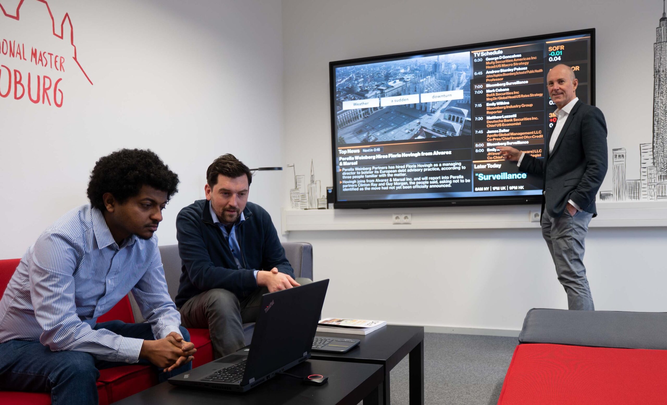 Three men are in a meeting room with a large screen displaying a presentation at Hochschule Coburg. One man stands near the screen, gesturing, while the other two sit on a red couch, one using a laptop. Wall graphics include "Hamburg," creating a professional and modern atmosphere.