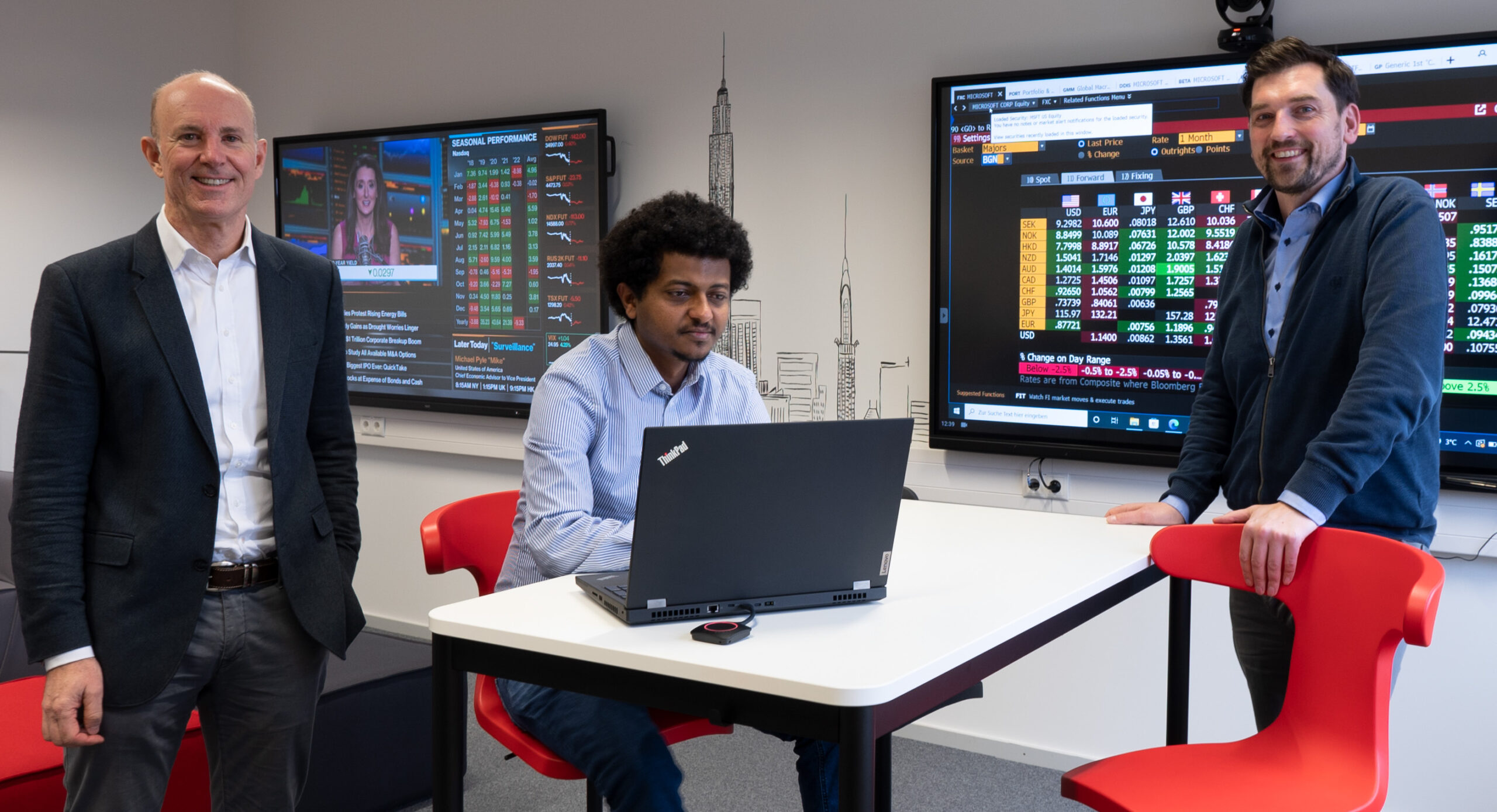 In a sleek, modern office at Hochschule Coburg, three men in business attire review financial data on large screens. One sits at a desk with a laptop while the other two stand beside him, smiling confidently at the camera.