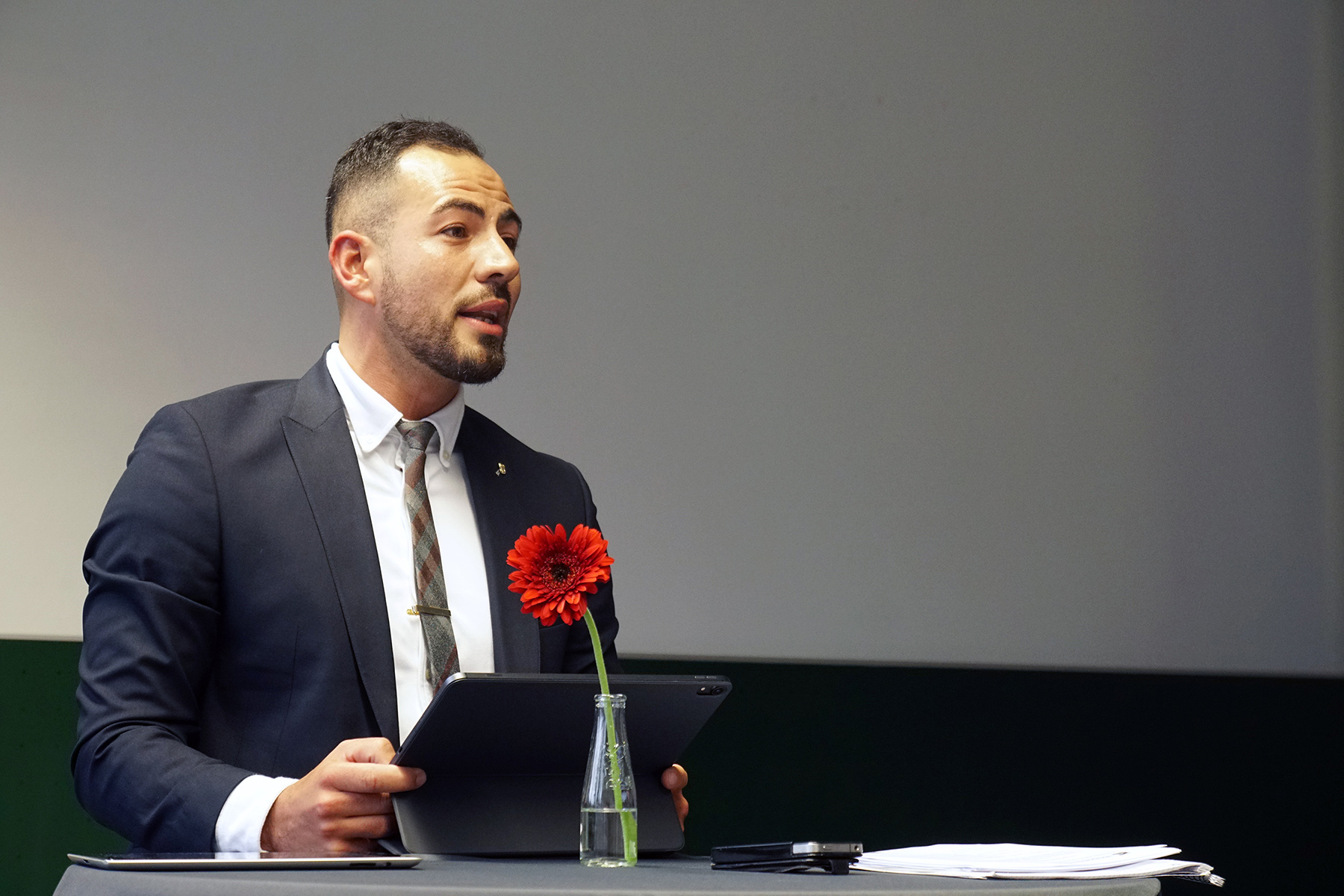 Ein Mann in Anzug und Krawatte steht hinter einem Tisch und hält ein Tablet mit dem Logo der Hochschule Coburg in der Hand. Auf dem Tisch liegen eine rote Blume in einer kleinen Glasflasche und einige Papiere. Im Hintergrund ist eine leere Wand.