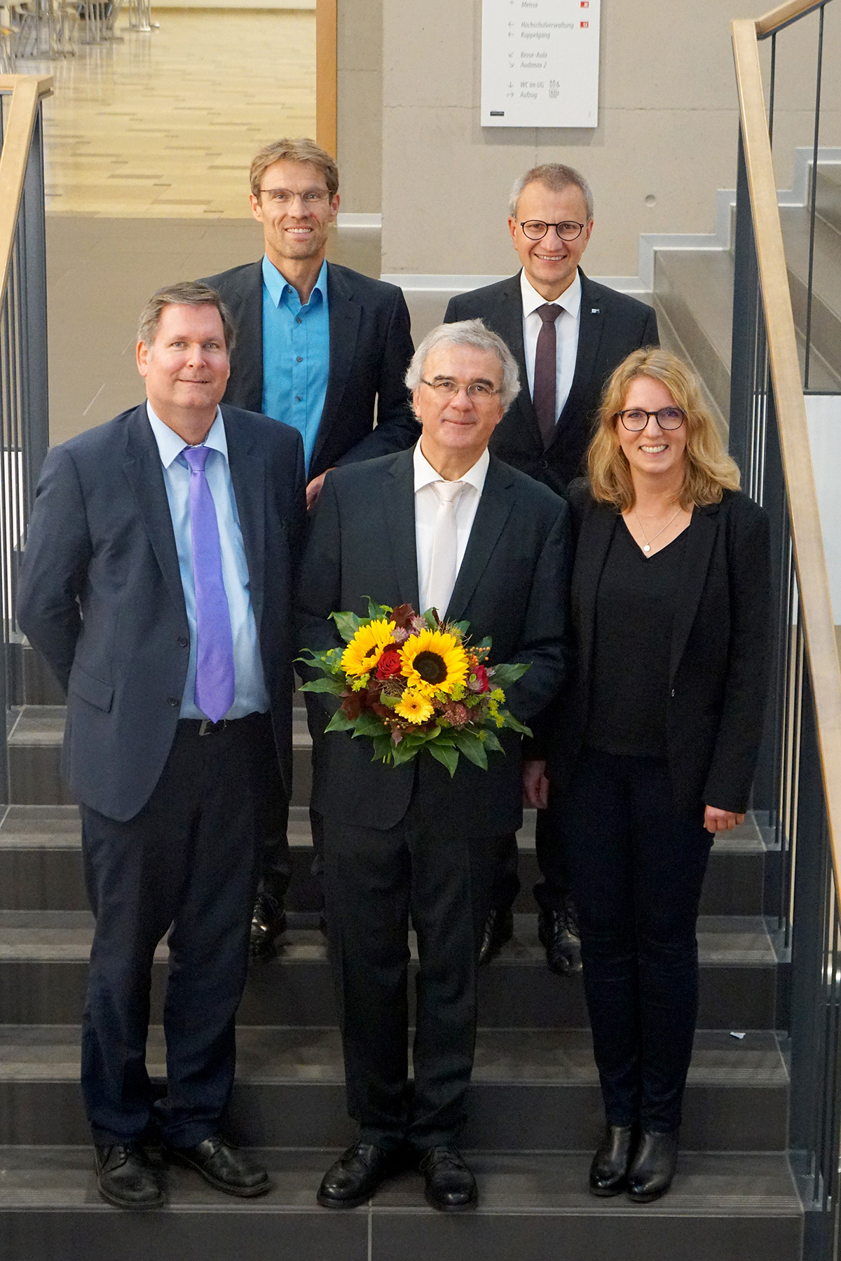 Eine Gruppe von fünf Personen, vier Männer und eine Frau, steht auf einer Treppe der Hochschule Coburg. Der Mann in der Mitte hält einen Strauß Sonnenblumen. Sie sind in formelle Kleidung gekleidet und lächeln alle herzlich in die Kamera.