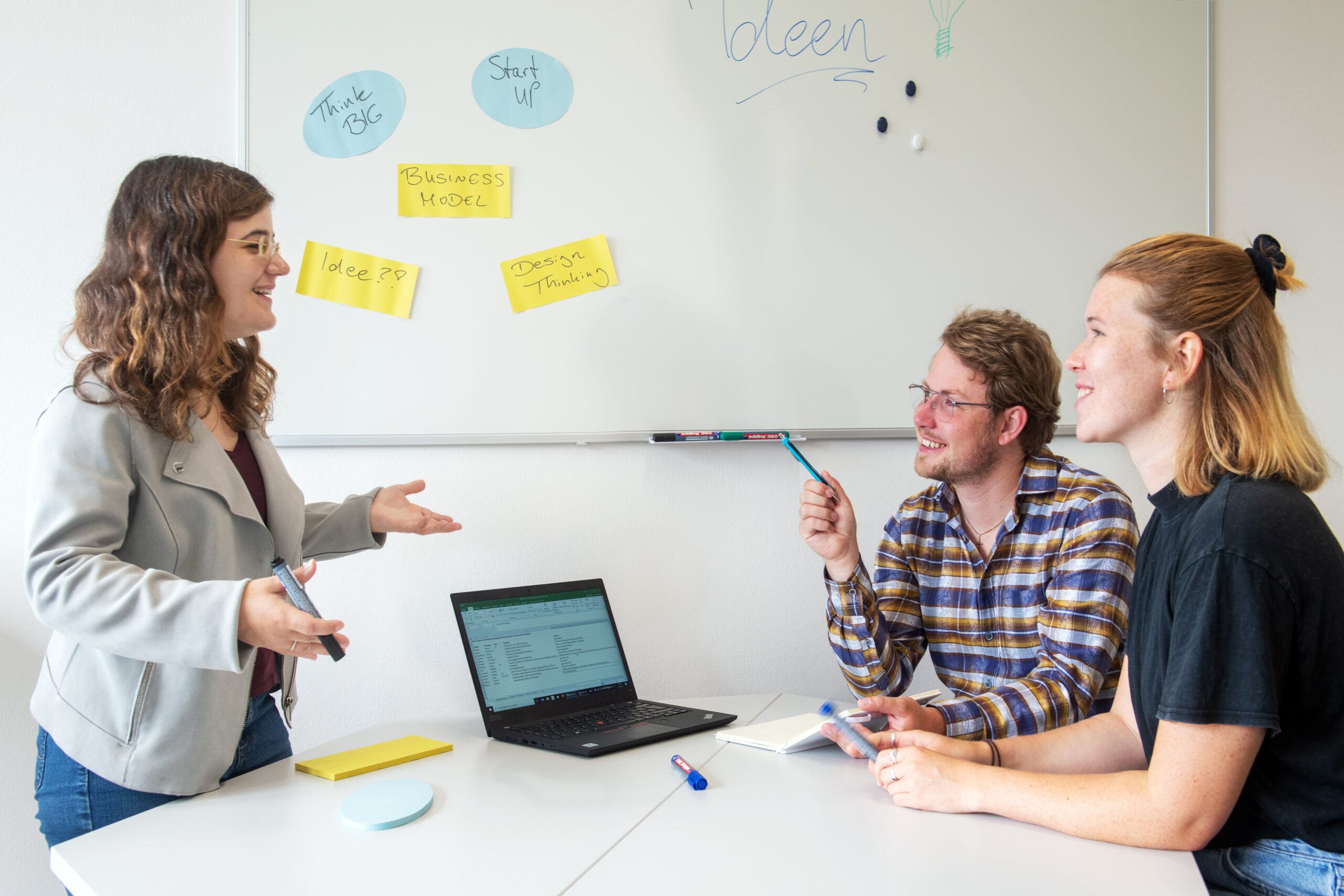 Drei Personen diskutieren in einem Büro über Ideen. Zwei sitzen mit einem Laptop an einem Tisch, die dritte steht und gestikuliert. Auf einem Whiteboard hinter ihnen hängen Haftnotizen zu Geschäftsmodellen und Startups, die innovative Konzepte der Hochschule Coburg reflektieren.