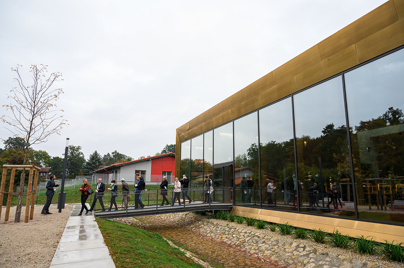 Eine Gruppe von Menschen schlendert einen Weg entlang und überquert eine kleine Brücke in Richtung des modernen Gebäudes der Hochschule Coburg mit seinen großen Glasfenstern. Das Gebäude ist wunderschön von üppigem Grün umgeben und hebt sich von einem klaren blauen Himmel ab.