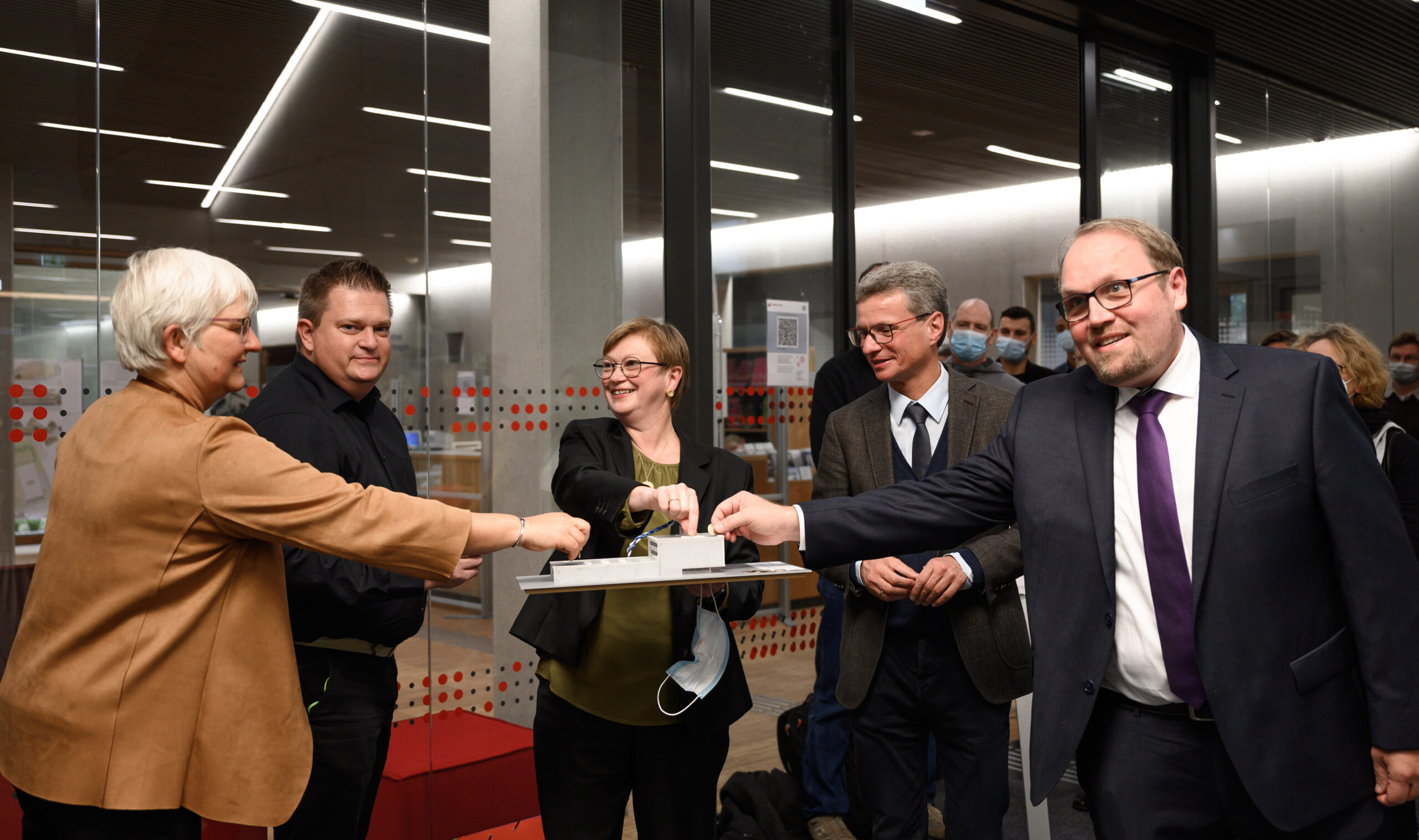 Eine Gruppe von sechs Personen in formeller Kleidung hat sich drinnen versammelt, lächelt und unterhält sich. Fünf von ihnen stoßen gemeinsam mit den Fäusten über einer Gedenktafel aneinander. Die Kulisse scheint ein Büro oder Konferenzraum mit Glaswänden, roten Akzenten und einer subtilen Anspielung auf den lebendigen Geist der Hochschule Coburg zu sein.