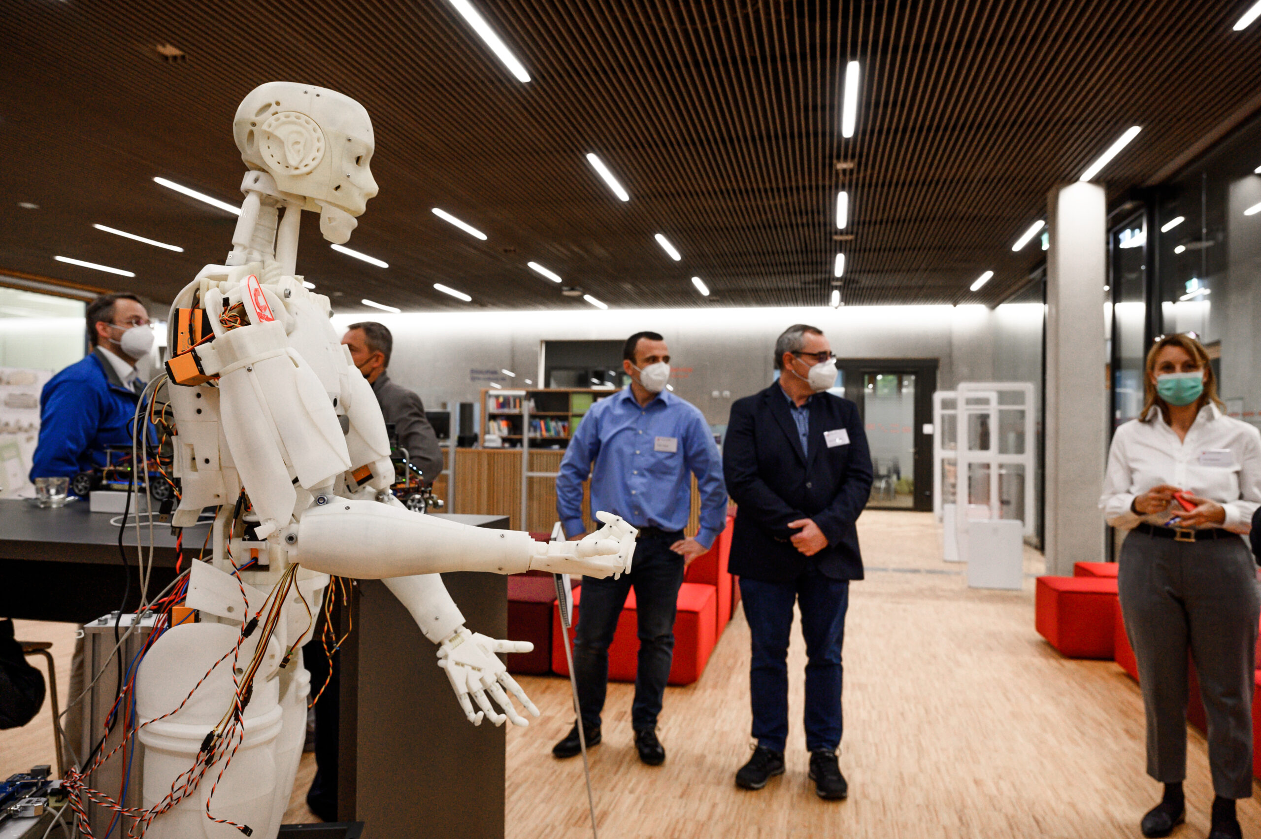 In einem modernen Raum der Hochschule Coburg ist ein humanoider Roboter mit freiliegenden Kabeln ausgestellt. Vier Personen stehen daneben und beobachten ihn mit Masken. Der Raum ist mit Holzboden, roten Sitzgelegenheiten und Regalen im Hintergrund ausgestattet.