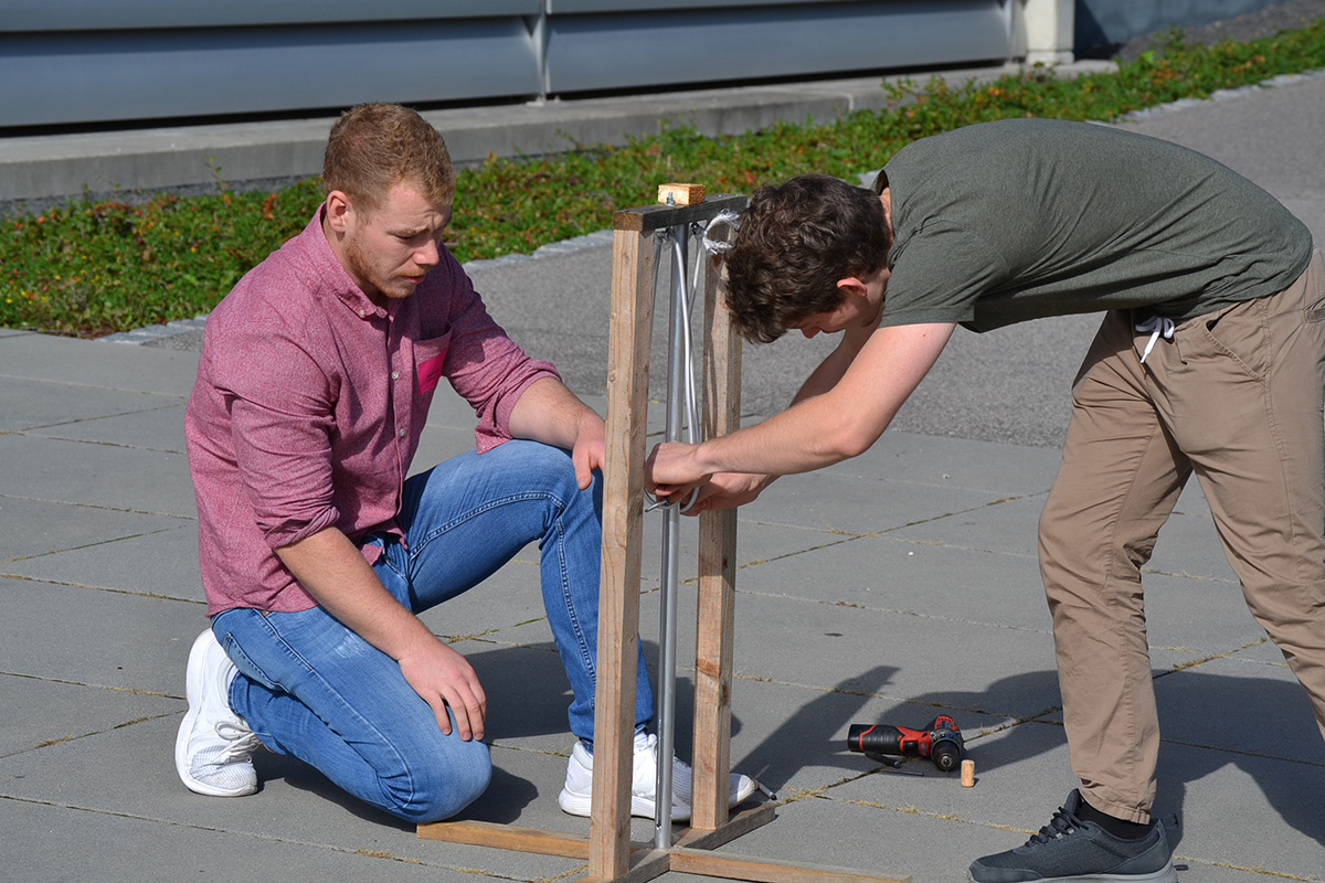 Zwei Männer bauen im Freien auf einer befestigten Fläche an der Hochschule Coburg ein Holzgerüst zusammen. Einer kniet und hält die Konstruktion fest, während der andere im Stehen ein Bauteil festzieht. Daneben liegt eine Akku-Bohrmaschine.