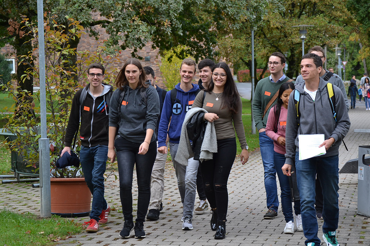 Eine Gruppe junger Erwachsener, bei denen es sich offenbar um Studierende der Hochschule Coburg handelt, geht auf einem gepflasterten Weg, der von Grün umgeben ist. Einige halten Papiere in den Händen und tragen legere Kleidung. Sie lächeln und scheinen in ein Gespräch vertieft zu sein.