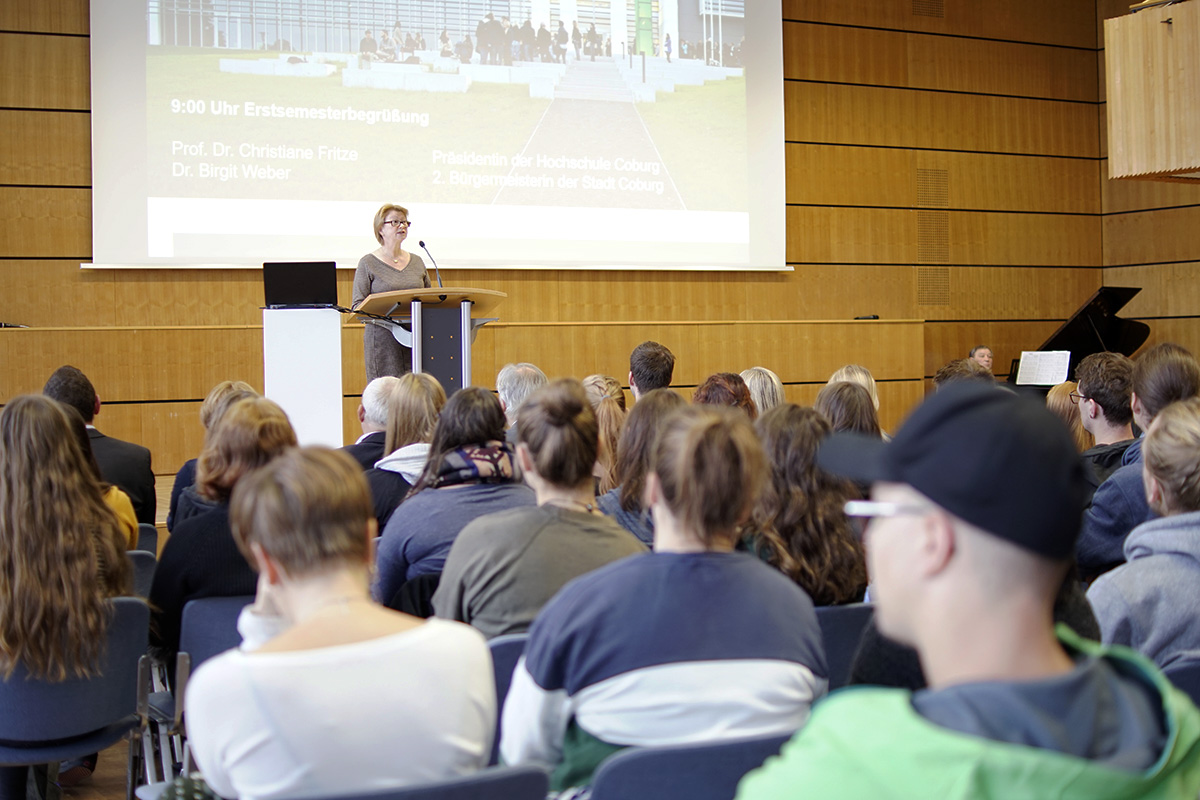 Eine Frau steht an einem Rednerpult im Hörsaal der Hochschule Coburg und spricht zu einem sitzenden Publikum. Hinter ihr läuft auf der Leinwand ein detaillierter Zeitplan. Das bunt gemischte Publikum hört aufmerksam zu und konzentriert sich ganz auf die spannende Präsentation, die vor ihnen liegt.