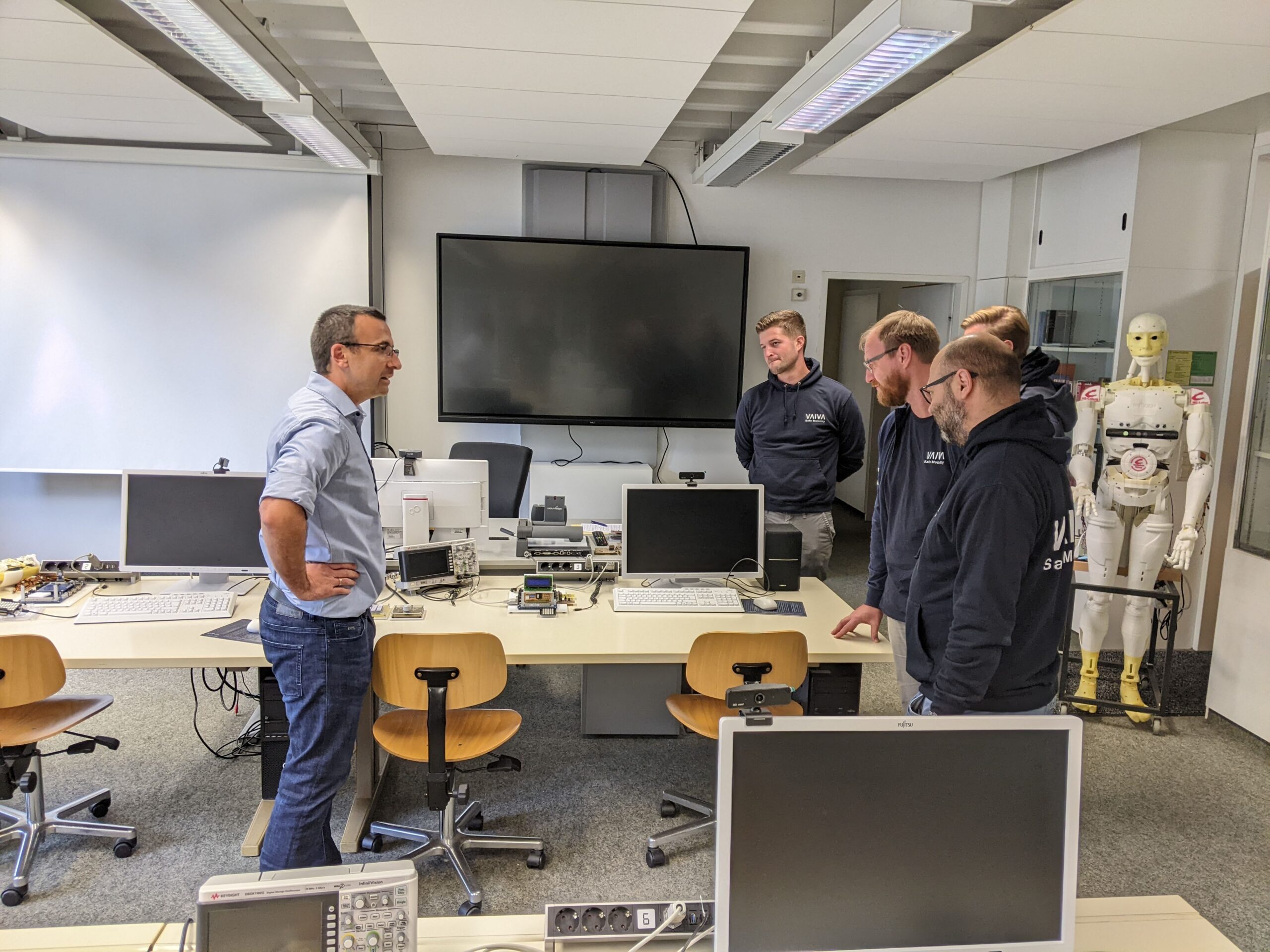 Eine Gruppe von fünf Personen steht in einem Laborraum der Hochschule Coburg. Im Hintergrund sind Computergeräte und ein humanoider Roboter zu sehen. Sie diskutieren, umgeben von Schreibtischen und Monitoren. An der Wand hängt ein großer Bildschirm, auf dem Daten angezeigt werden.