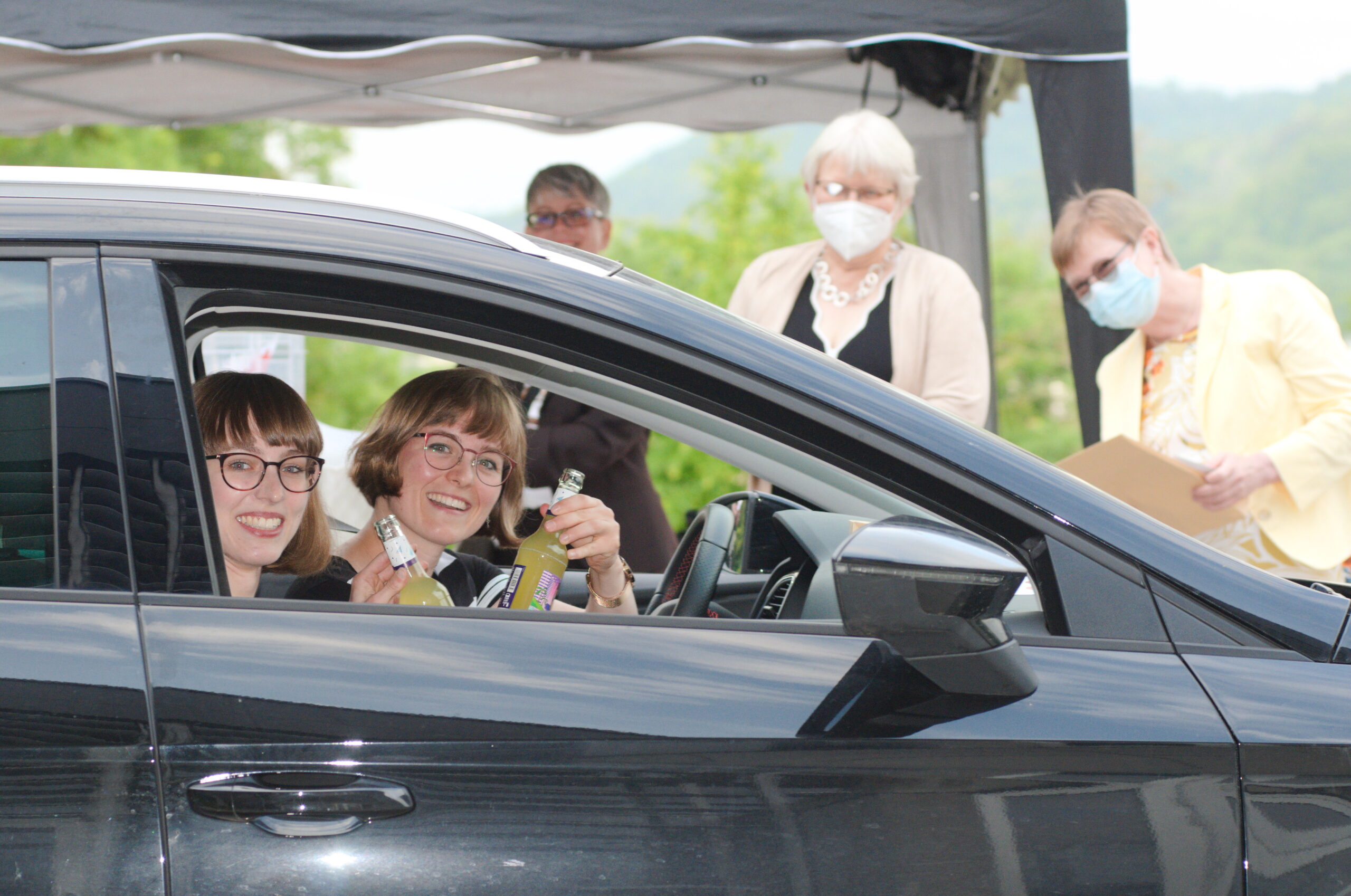 Zwei Menschen sitzen in einem Auto, lächeln und halten kleine Gegenstände in den Händen. Draußen steht daneben eine Gruppe der Hochschule Coburg mit Masken, eine Person hält einen Ordner in der Hand. Im Hintergrund ist ein Baldachin zu sehen.