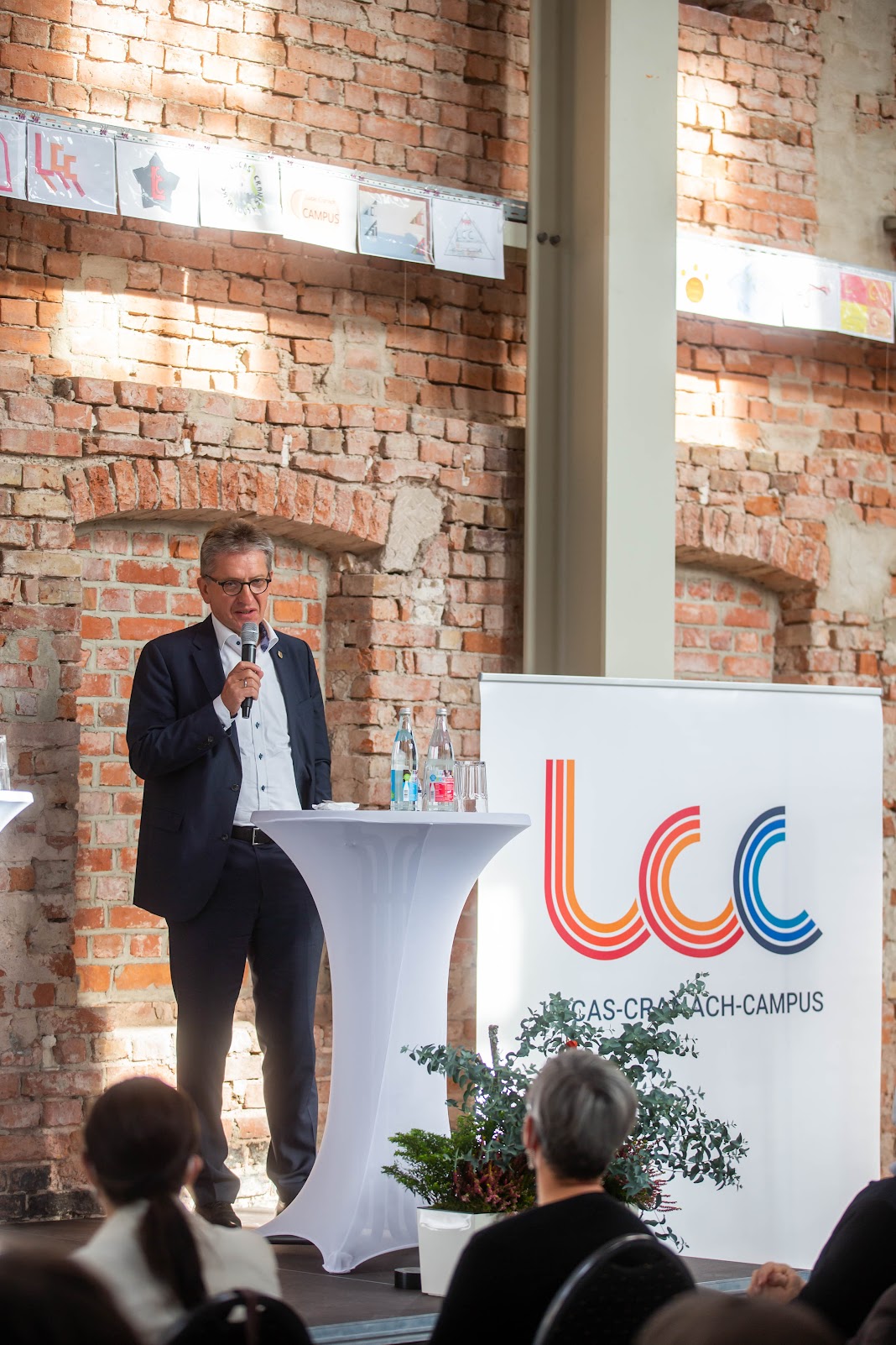 Ein Mann im Anzug spricht in ein Mikrofon an einem Podium, das mit den Logos „LCC“ und „CAS-Campus“ geschmückt ist und an Veranstaltungen an der Hochschule Coburg erinnert. Die Szene besteht aus Backsteinmauern und einer Kulisse, während ein kleines Publikum aufmerksam zuhört. Neben dem Podium steht eine dekorative Pflanze.