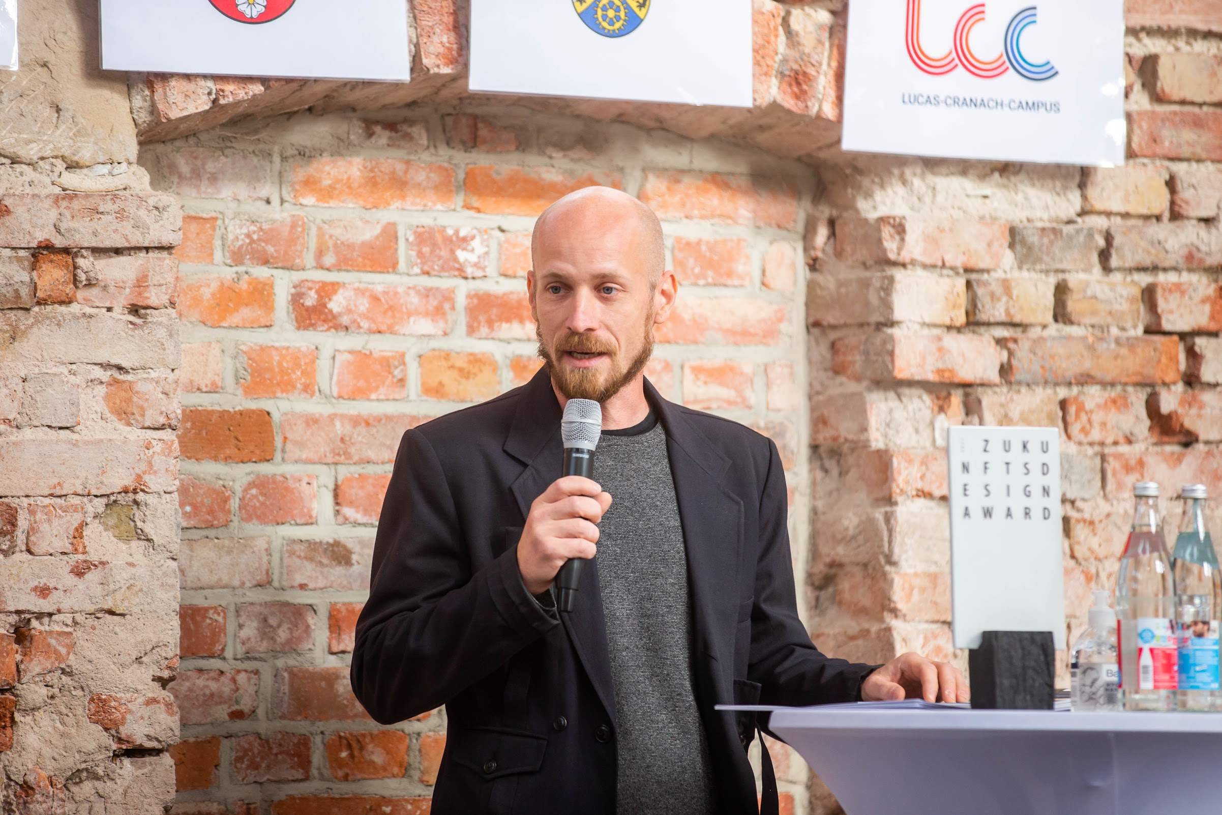 Ein Mann mit Bart, schwarzer Jacke und grauem Hemd spricht in ein Mikrofon. Er steht vor einer Backsteinwand, über ihm sind Logos der Hochschule Coburg zu sehen, neben ihm ein kleiner Tisch mit Gläsern und Flaschen.