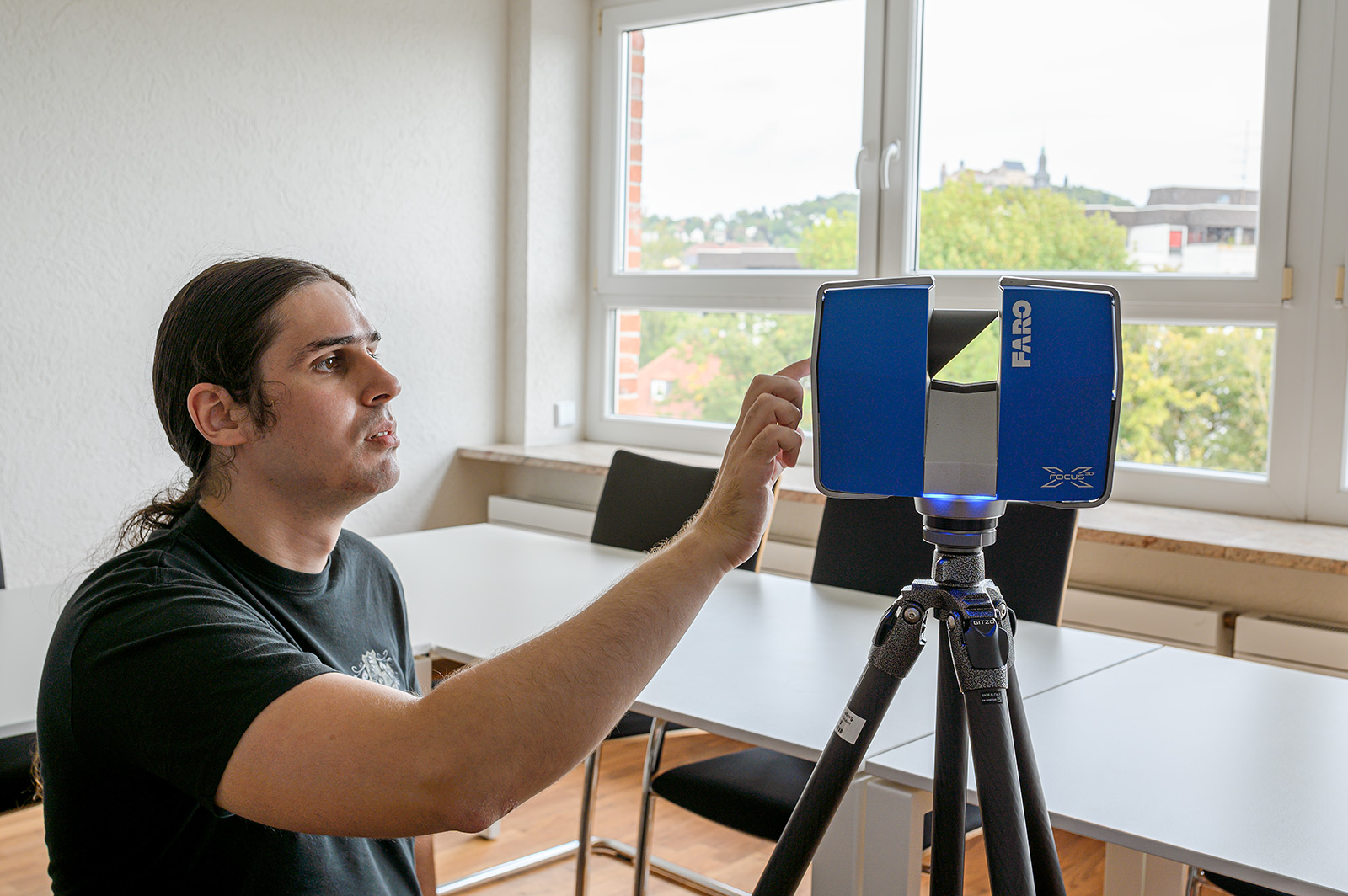 Eine Person mit langen Haaren bedient einen FARO-Laserscanner auf einem Stativ in einem Raum voller Tische und Stühle. Durch das Fenster dahinter bietet sich ein malerischer Blick auf Bäume und entfernte Gebäude, der den Innovationsgeist der Hochschule Coburg einfängt.