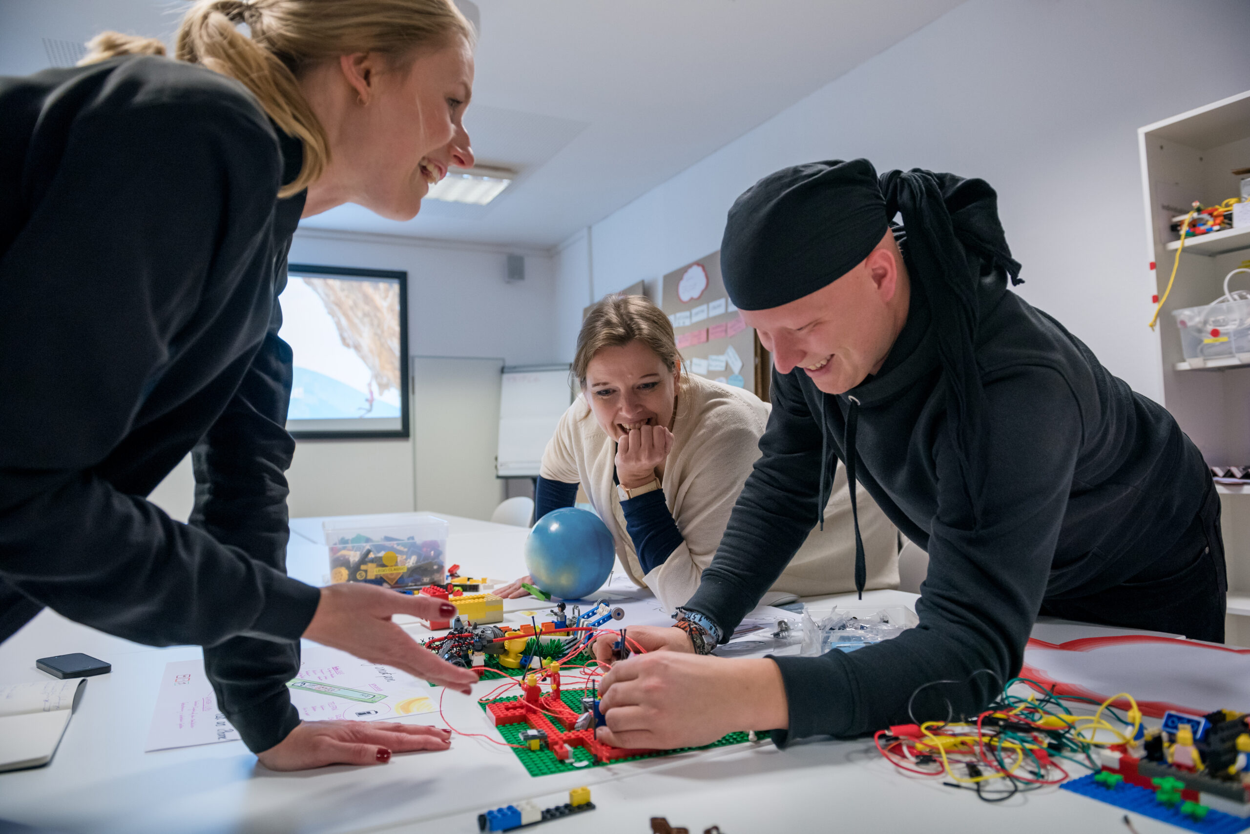 An einem Tisch voller bunter Bauklötze und Drähte arbeiten drei Personen in einem innovativen Geist zusammen, der an die Hochschule Coburg erinnert. Einer beugt sich lächelnd vor, während ein anderer konzentriert die Teile anordnet. Im Hintergrund leuchtet eine Projektionsleinwand und suggeriert eine kreative oder pädagogische Umgebung.