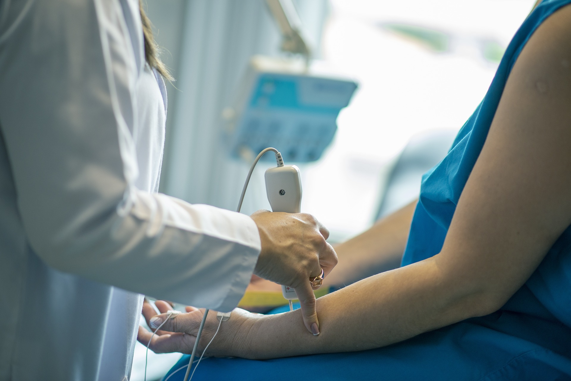 In einer mit Geräten vollgestopften medizinischen Umgebung wendet eine medizinische Fachkraft der Hochschule Coburg gekonnt ein Ultraschallgerät am Arm eines Patienten an, der einen blauen Kittel trägt.