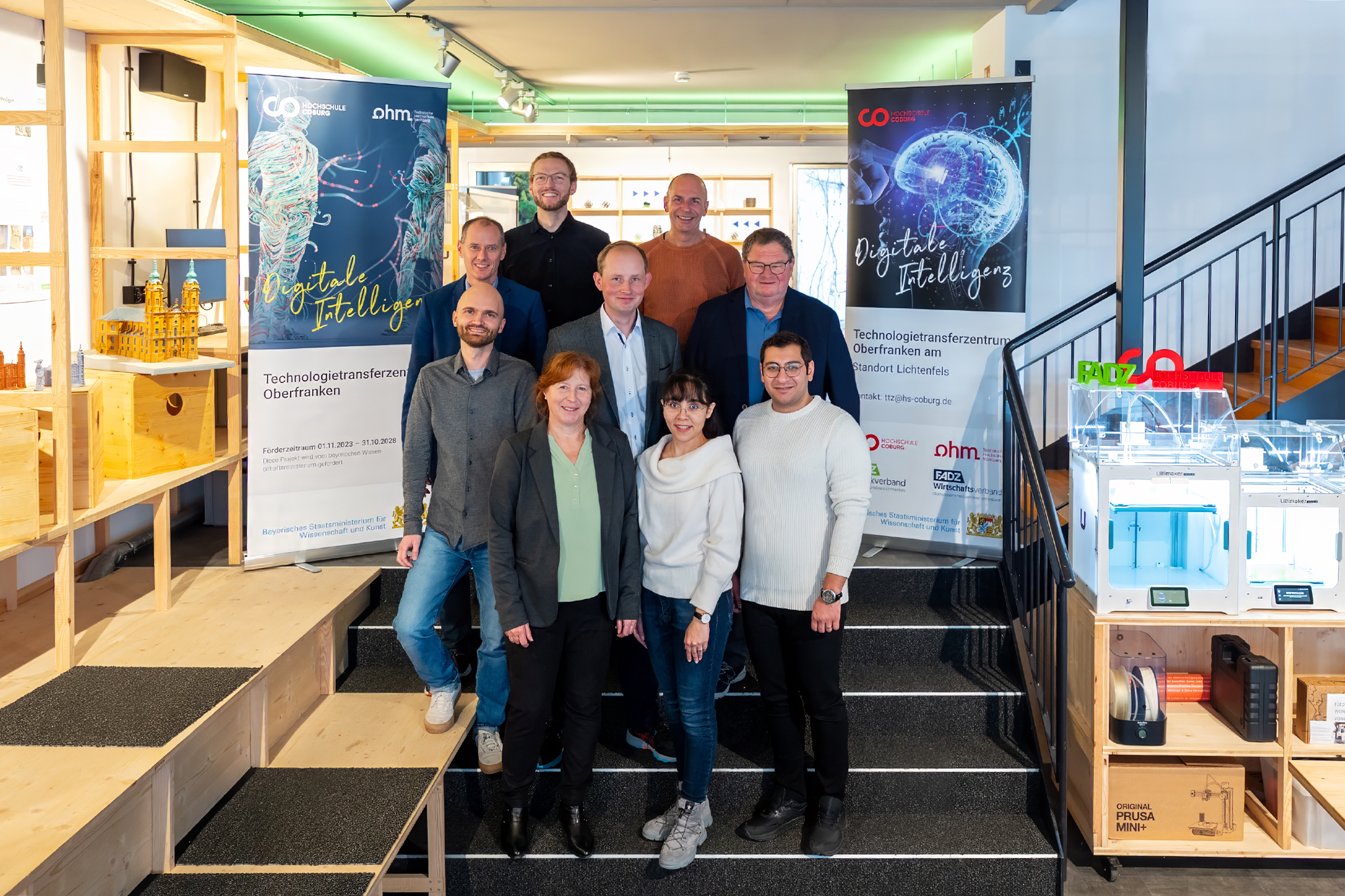 Eine Gruppe von neun Personen posiert auf einer Treppe im Technologiezentrum der Hochschule Coburg. Im Hintergrund sind zwei Banner mit dem Text „Digitale Intelligenz“ zu sehen. Im Raum sind verschiedene technische Geräte zu sehen.