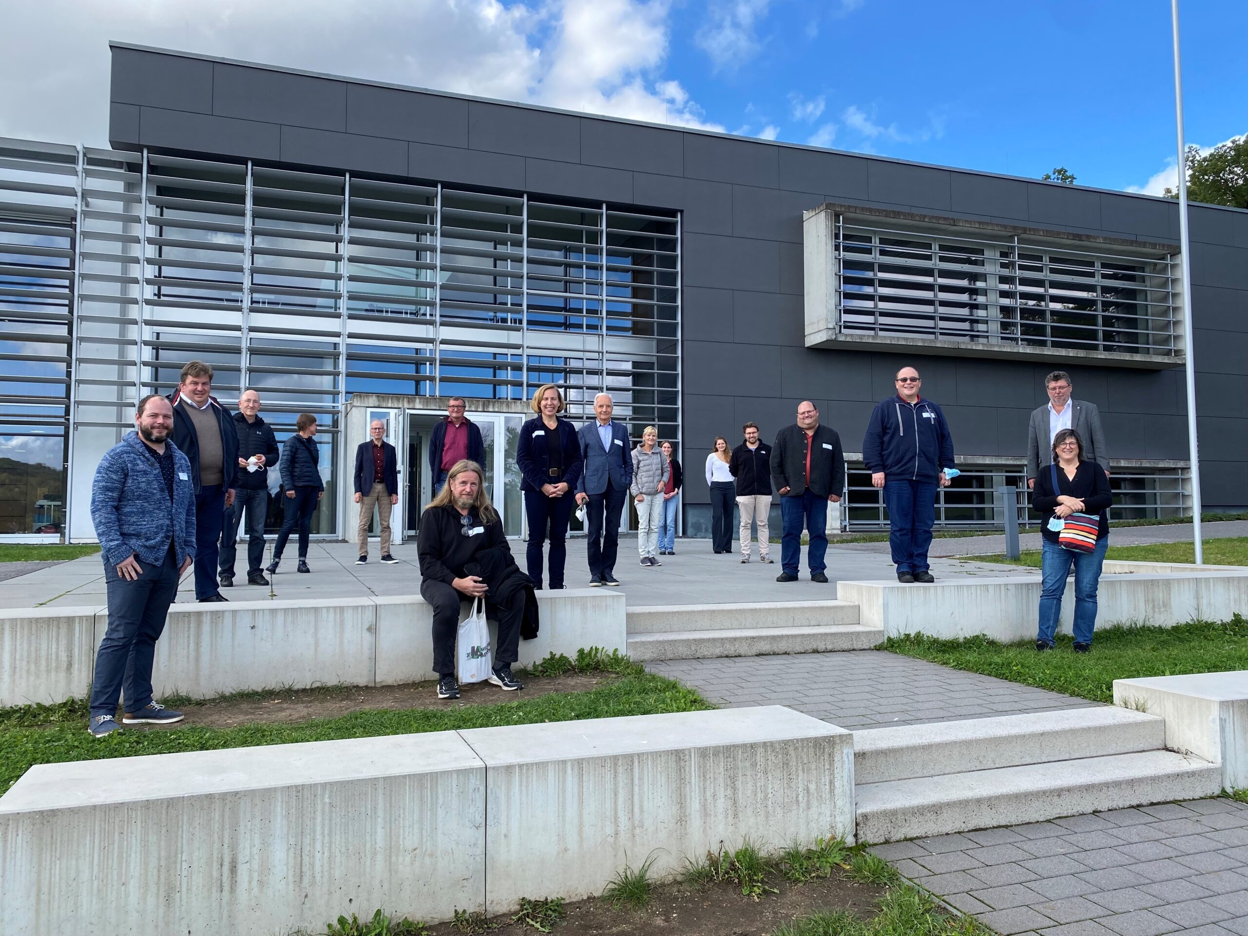 Eine Gruppe von Menschen steht und sitzt auf den Stufen vor dem modernen Gebäude der Hochschule Coburg mit seinen großen Fenstern und grauen Paneelen. Der Himmel ist teilweise bewölkt. Sie sind leger gekleidet und lächeln in die Kamera. Sie fangen einen Moment der Wärme und Gemeinschaft ein.