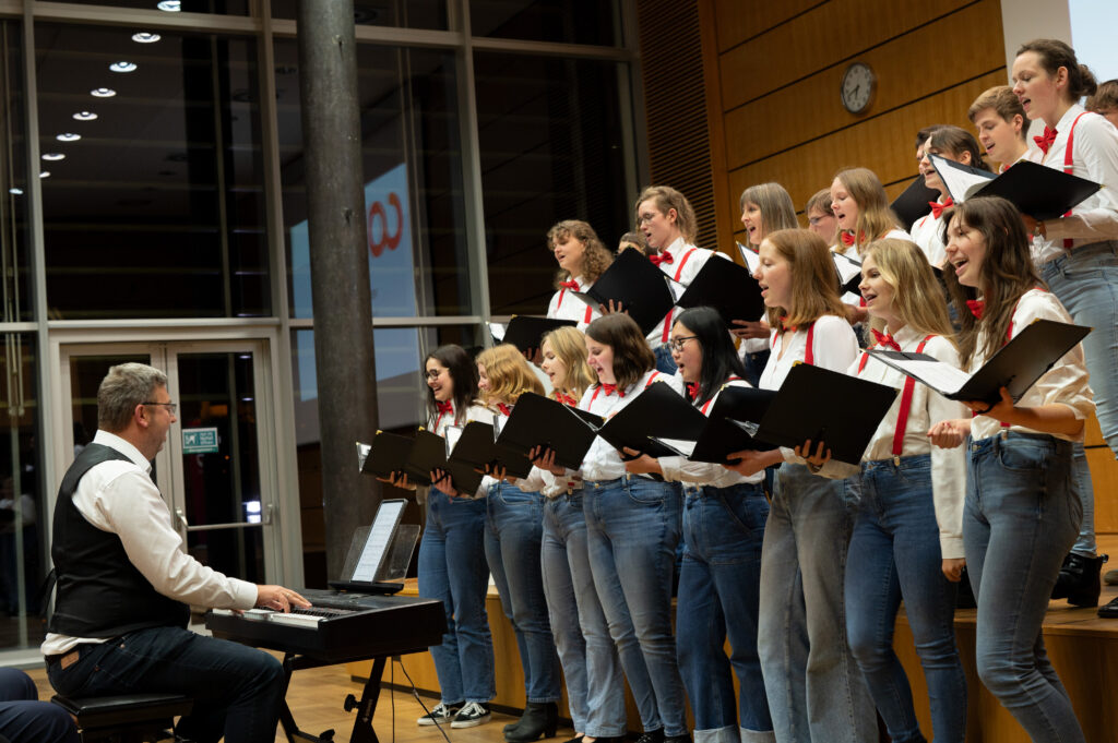 In an indoor hall with glass walls, the talented choir of Hochschule Coburg’s women passionately sings in white shirts and jeans, each adorned with a red tie and holding black folders. They are artfully directed by a man in a white shirt and black vest playing the keyboard.