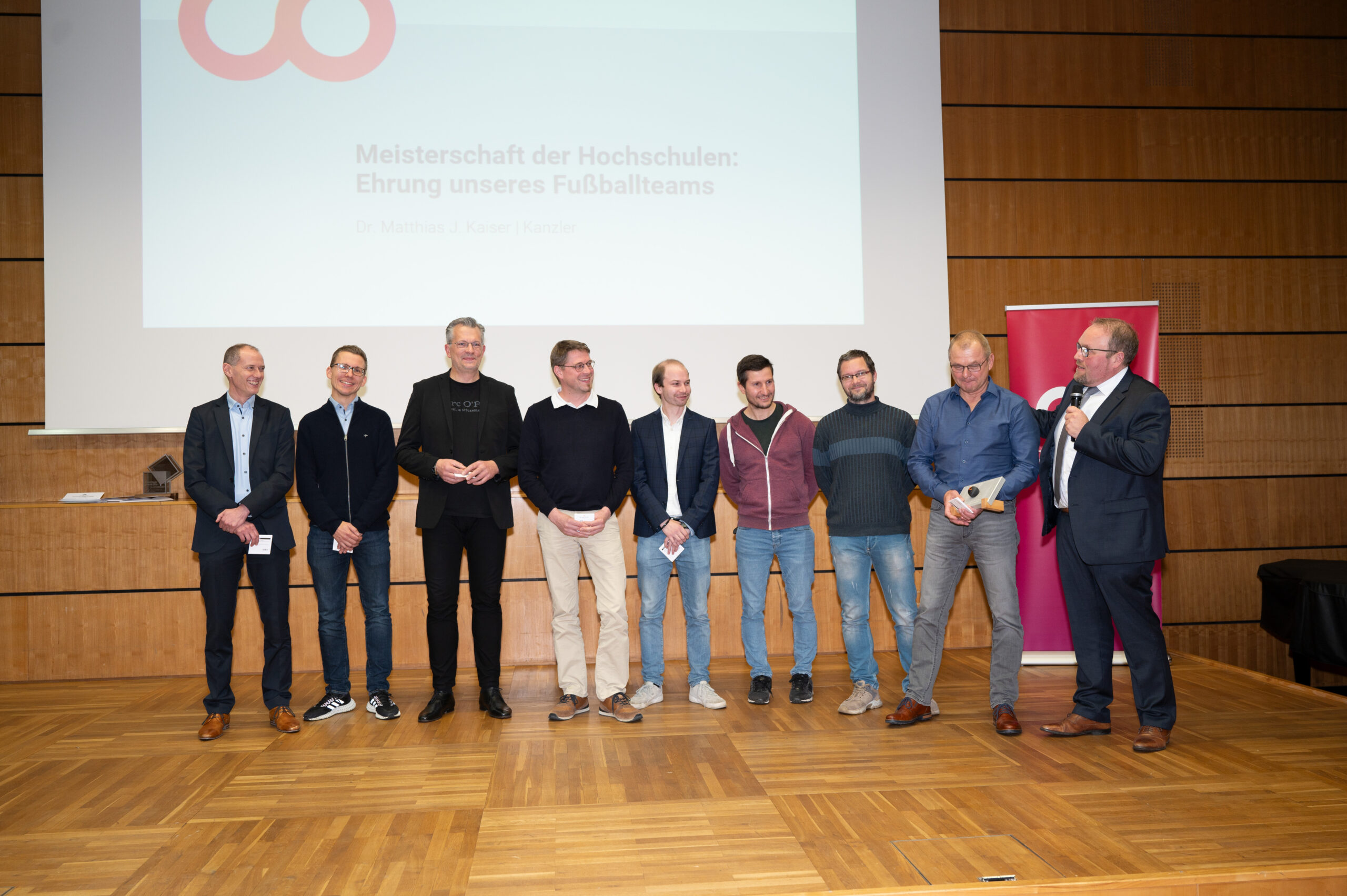 Eine Gruppe von neun Männern steht während einer Preisverleihung an der Hochschule Coburg in einer Reihe auf einer Bühne. Ein Mann ganz rechts spricht in ein Mikrofon, während auf einem Bildschirm hinter ihnen ein Text zu Ehren einer Fußballmannschaft angezeigt wird. Die Kulisse scheint eine formelle Veranstaltung in einer Halle zu sein.