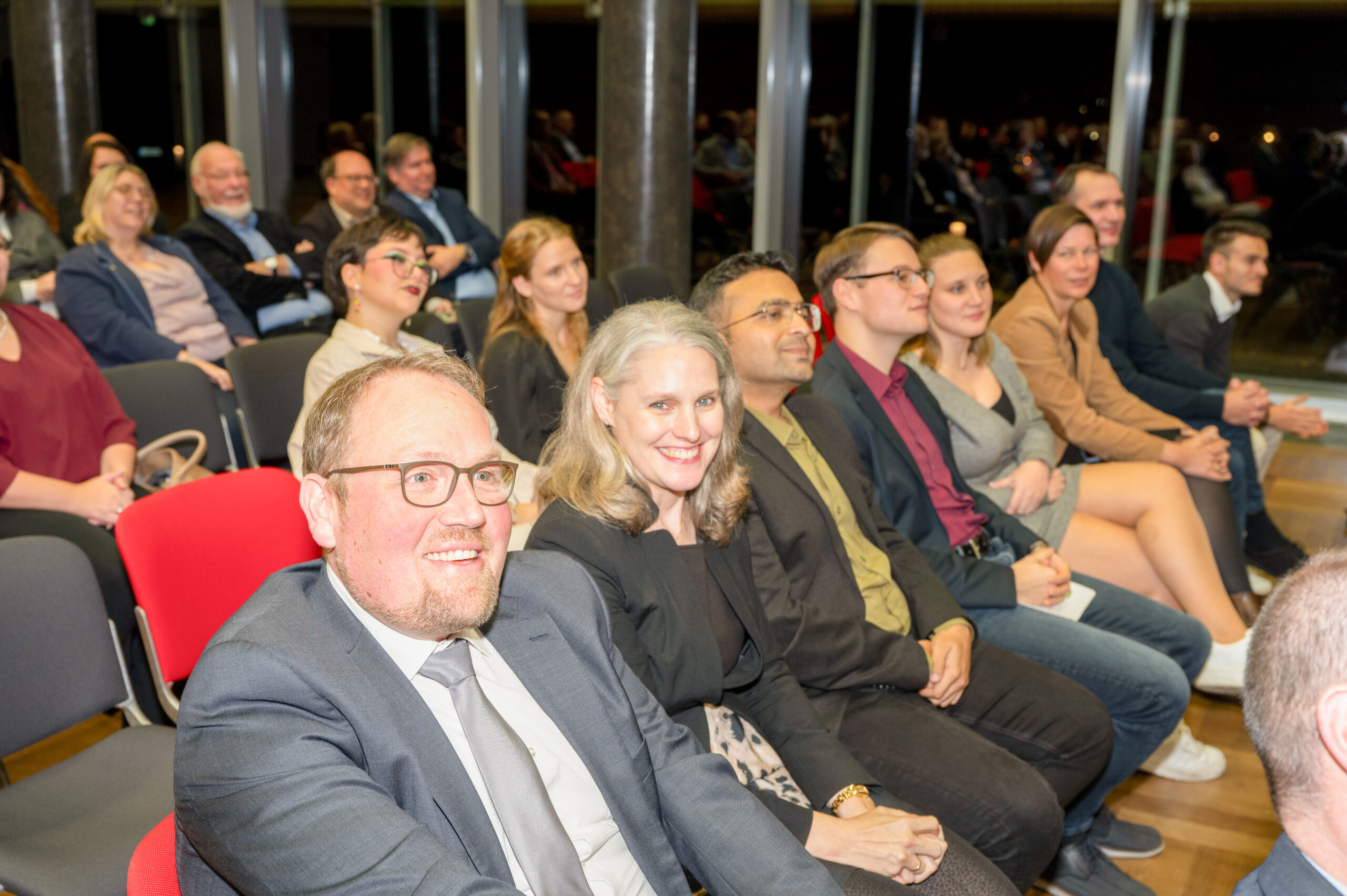 Bei einer Indoor-Veranstaltung der Hochschule Coburg sitzt eine Gruppe von Menschen in Stuhlreihen. Sie sind formell gekleidet, wirken engagiert und lächeln, während sie nach vorne blicken. Die Umgebung lässt auf eine Versammlung wie ein Seminar, eine Vorlesung oder eine Konferenz schließen.