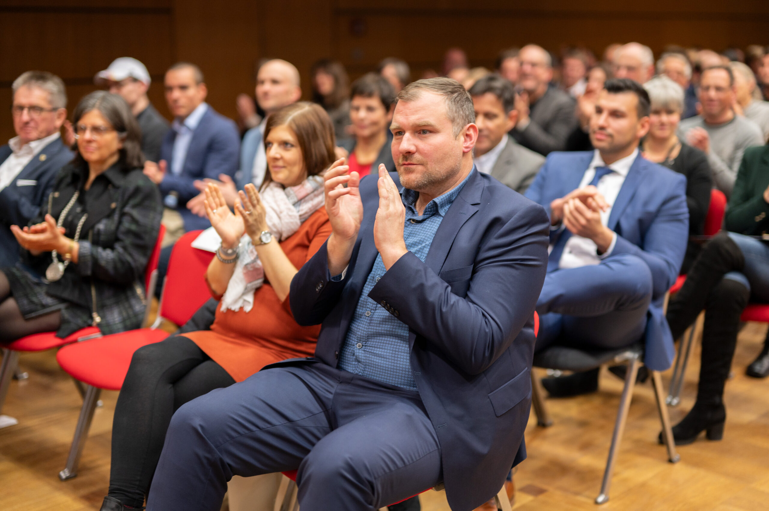 Eine Gruppe von Menschen sitzt auf roten Stühlen in einem großen Raum der Hochschule Coburg und verfolgt aufmerksam eine Veranstaltung. Der Mann im Vordergrund klatscht. Er trägt einen blauen Anzug. Andere in Freizeit- und Abendkleidung klatschen ebenfalls. Den Hintergrund bilden ein Holzboden und eine Holzwand.