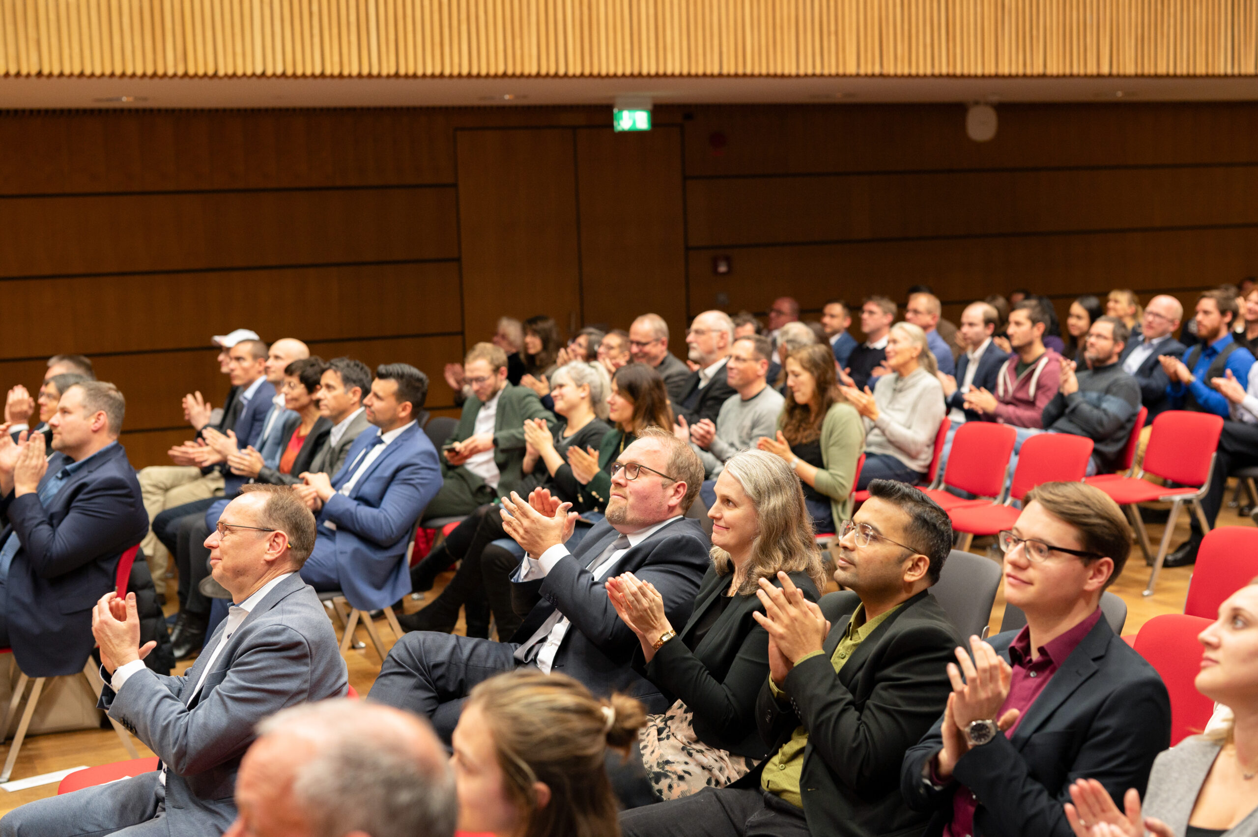 In einem großen Konferenzraum der Hochschule Coburg sitzt ein Publikum und applaudiert. Die bunt gemischte Gruppe, die sich in Alter und ethnischer Zugehörigkeit unterscheidet, sitzt auf roten Stühlen, einige tragen Anzüge, andere sind leger gekleidet, alle umgeben von Holzwänden, die eine formelle Umgebung suggerieren.